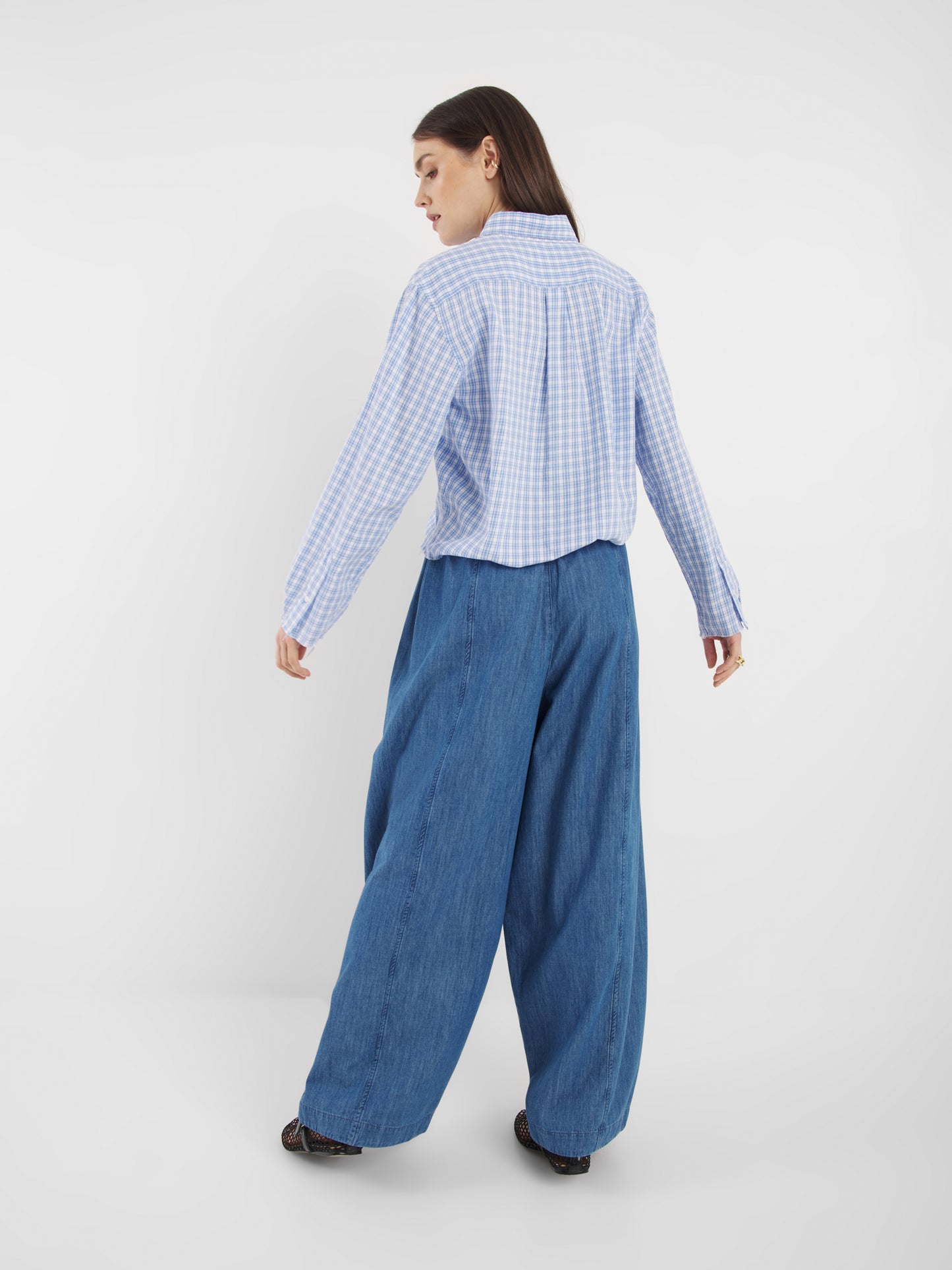 Eine Frau mit glattem, offenem Haar steht vor einem schlichten weißen Hintergrund und trägt das langärmelige Hemd Blouse Casual Check Blue White Check und eine weit geschnittene Blue Jeans.