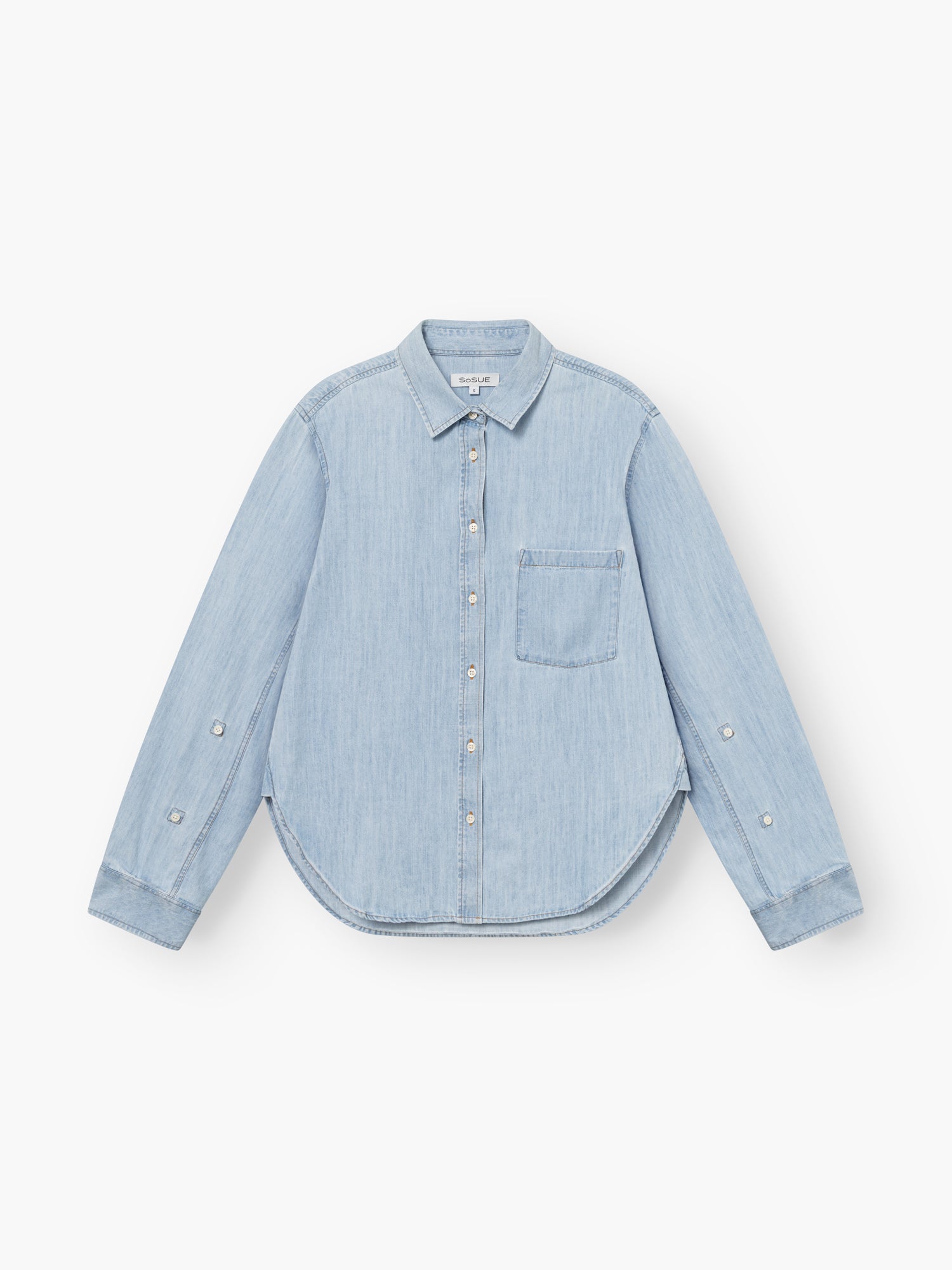 Bluse Classic Denim in Hellblau mit langen Ärmeln, einer Brusttasche, tailliertem Schnitt und geschwungenem Saum, flach auf weißem Grund gelegt.