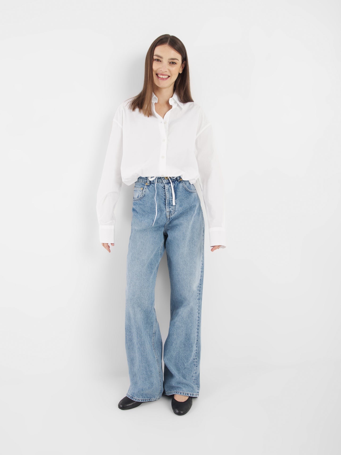Eine Frau steht vor einem weißen Hintergrund und lächelt. Sie trägt die Bluse Draw String White zu einer hochgeschlossenen, locker sitzenden Bluejeans mit gebundener Taille und schwarzen Schuhen.