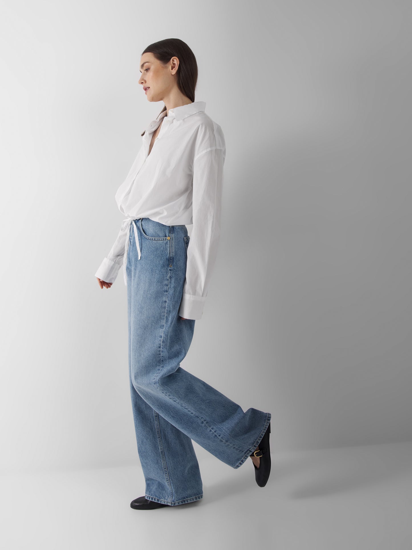 Eine Frau steht seitlich vor einem unifarbenen Hintergrund. Sie trägt die Bluse Draw String White, hellblaue Jeans mit hoher Taille und weiten Beinen und schwarze Schuhe, wobei sie einen Fuß leicht anhebt, um einen entspannten Sommerbluse Damen-Look zu erzielen.