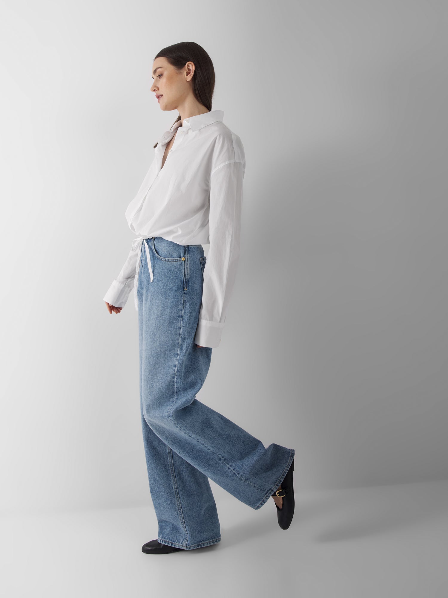 Eine Frau steht seitlich vor einem unifarbenen Hintergrund. Sie trägt die Bluse Draw String White, hellblaue Jeans mit hoher Taille und weiten Beinen und schwarze Schuhe, wobei sie einen Fuß leicht anhebt, um einen entspannten Sommerbluse Damen-Look zu erzielen.