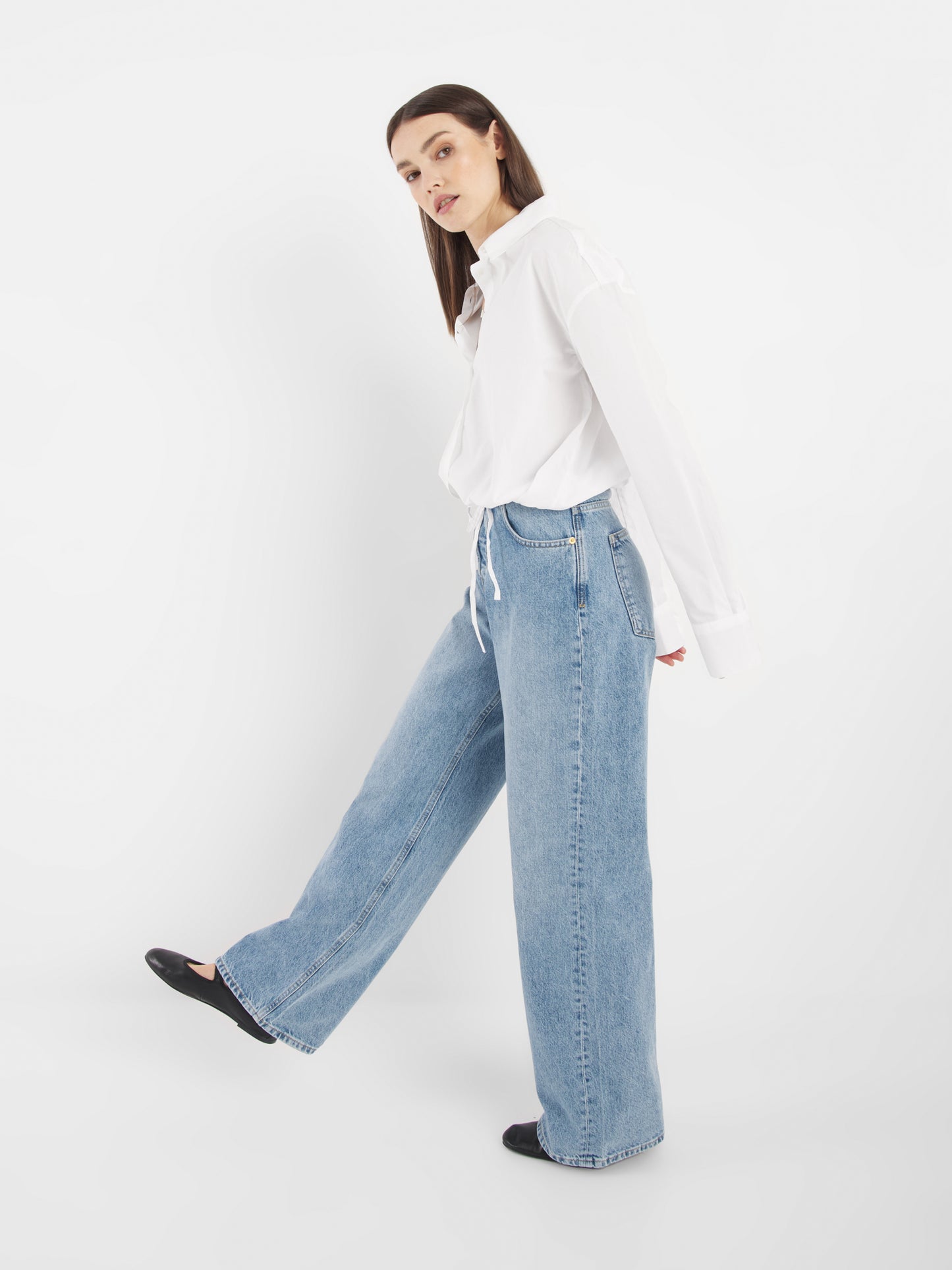 Eine Frau steht vor einem weißen Hintergrund und trägt die Bluse Draw String White und eine hoch sitzende, weit geschnittene Bluejeans. Sie blickt nach vorne, ein Bein leicht angehoben, und nimmt eine lässige Pose in diesem sommerlichen Look ein.