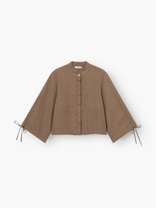 Die Bluse Mary Linen Soft Brown ist eine braune, locker sitzende Leinenjacke mit Mandarinenkragen, Knopfverschluss vorne, zwei Taschen vorne und weiten Ärmeln, die an den Handgelenken gebunden werden, auf einem schlichten weißen Hintergrund.
