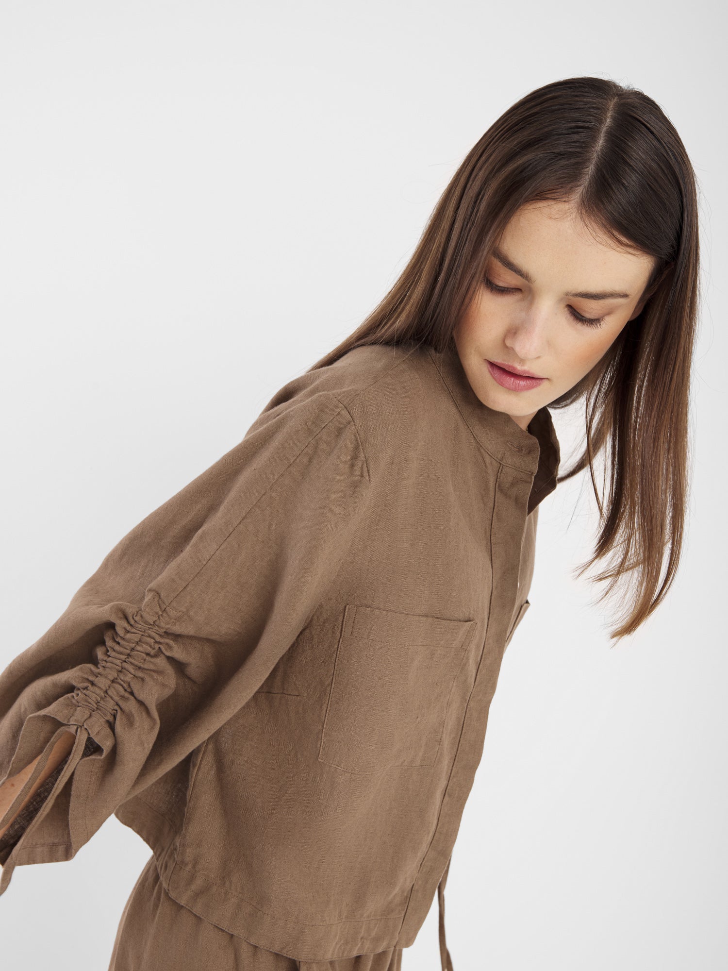 Eine junge Frau mit glattem braunem Haar blickt an sich herunter, während sie die Bluse Mary Linen Soft Brown trägt, eine locker sitzende Bluse mit gerafften Ärmeln, die vor einem schlichten weißen Hintergrund steht.