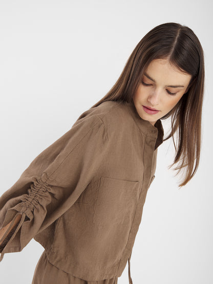 Eine junge Frau mit glattem braunem Haar blickt an sich herunter, während sie die Bluse Mary Linen Soft Brown trägt, eine locker sitzende Bluse mit gerafften Ärmeln, die vor einem schlichten weißen Hintergrund steht.
