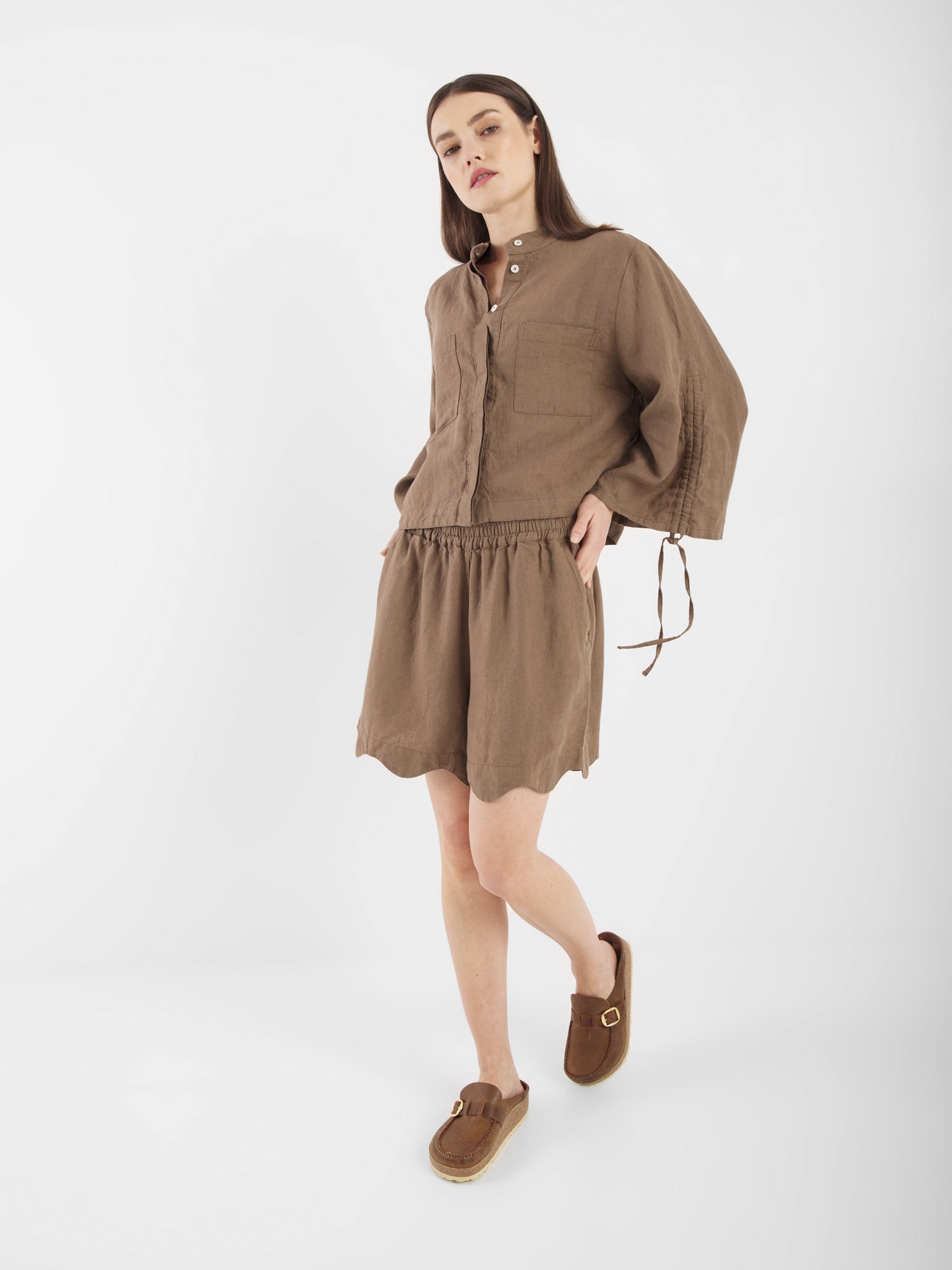Eine Frau steht vor einem weißen Hintergrund und trägt die Bluse Mary Linen Soft Brown mit Knöpfen, einen passenden braunen Rock und braune Slipper. Sie hat glattes braunes Haar und einen neutralen Gesichtsausdruck.