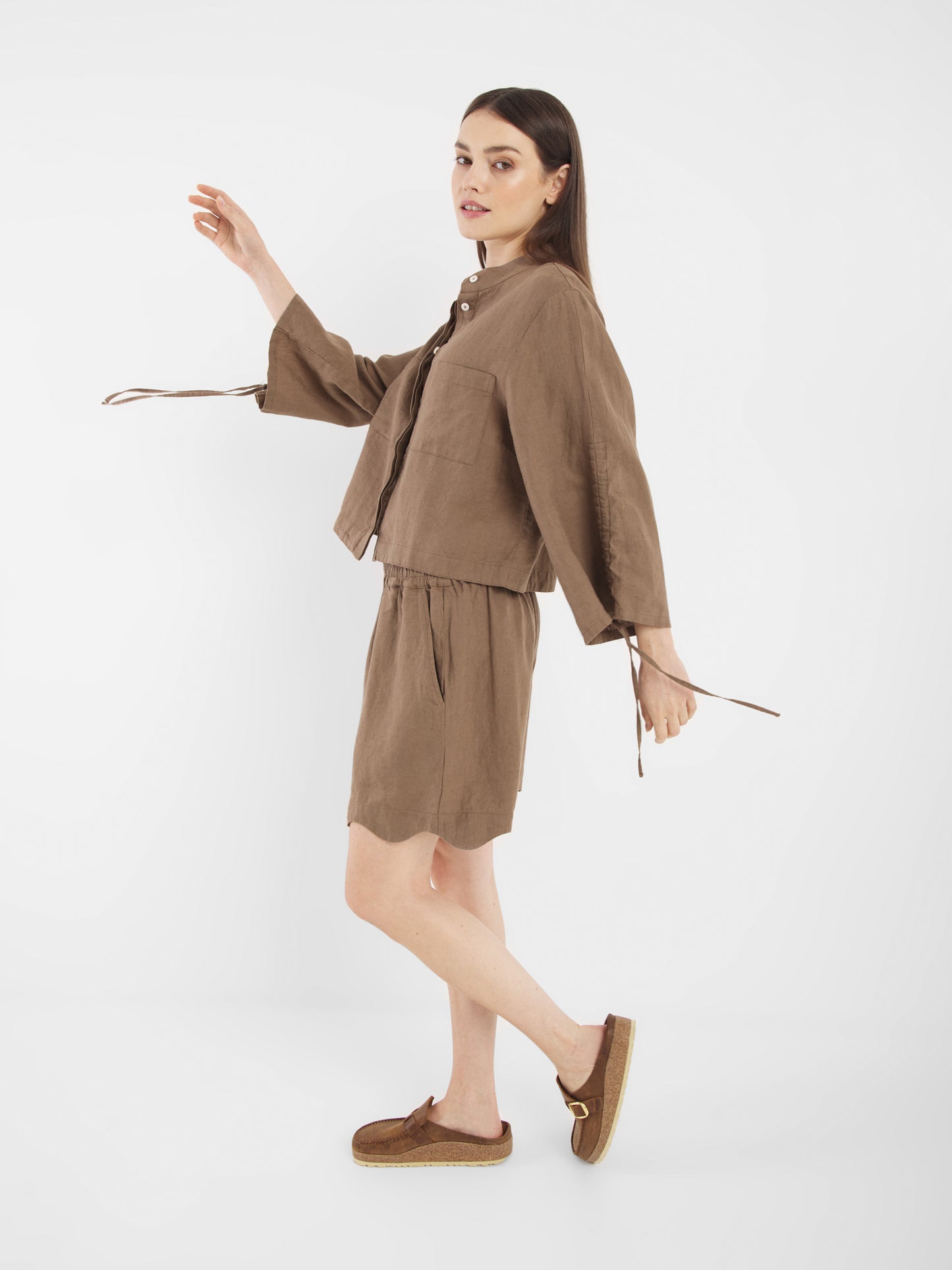 Eine Frau modelliert die Bluse Mary Linen Soft Brown mit einem passenden Rock und braunen Schuhen. Sie steht seitlich, ein Bein leicht angehoben, und schaut in die Kamera vor einem schlichten weißen Hintergrund.
