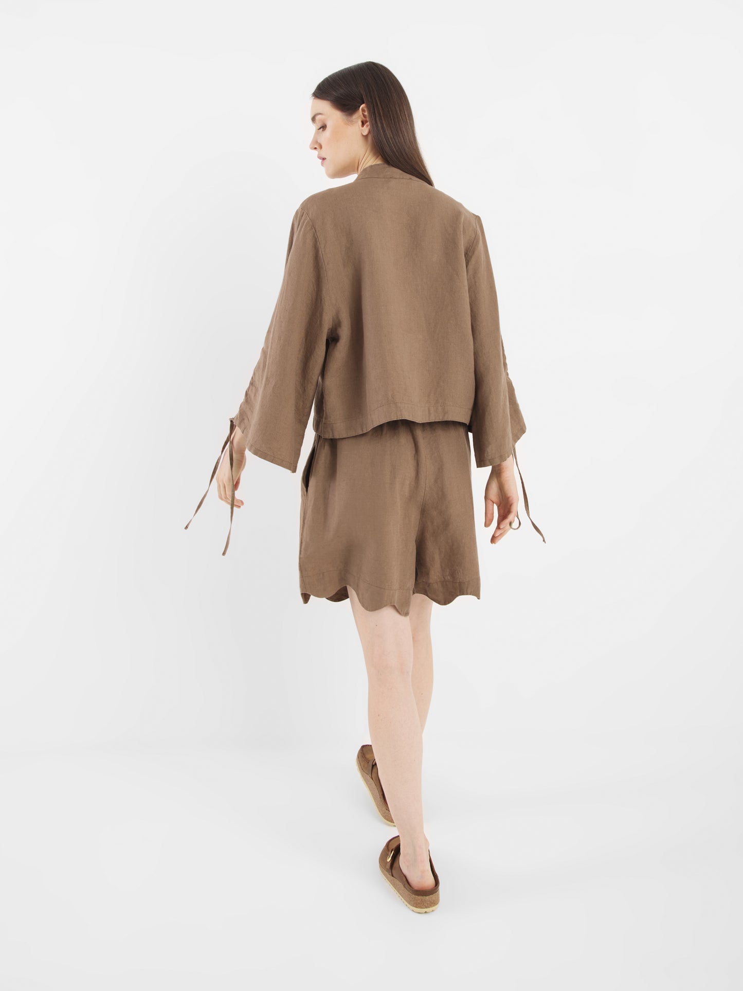 Eine Frau mit langen braunen Haaren blickt nach vorne. Sie trägt die Shorts Wave Soft Brown mit einer passenden lockeren Jacke und flachen braunen Schuhen vor einem schlichten weißen Hintergrund.