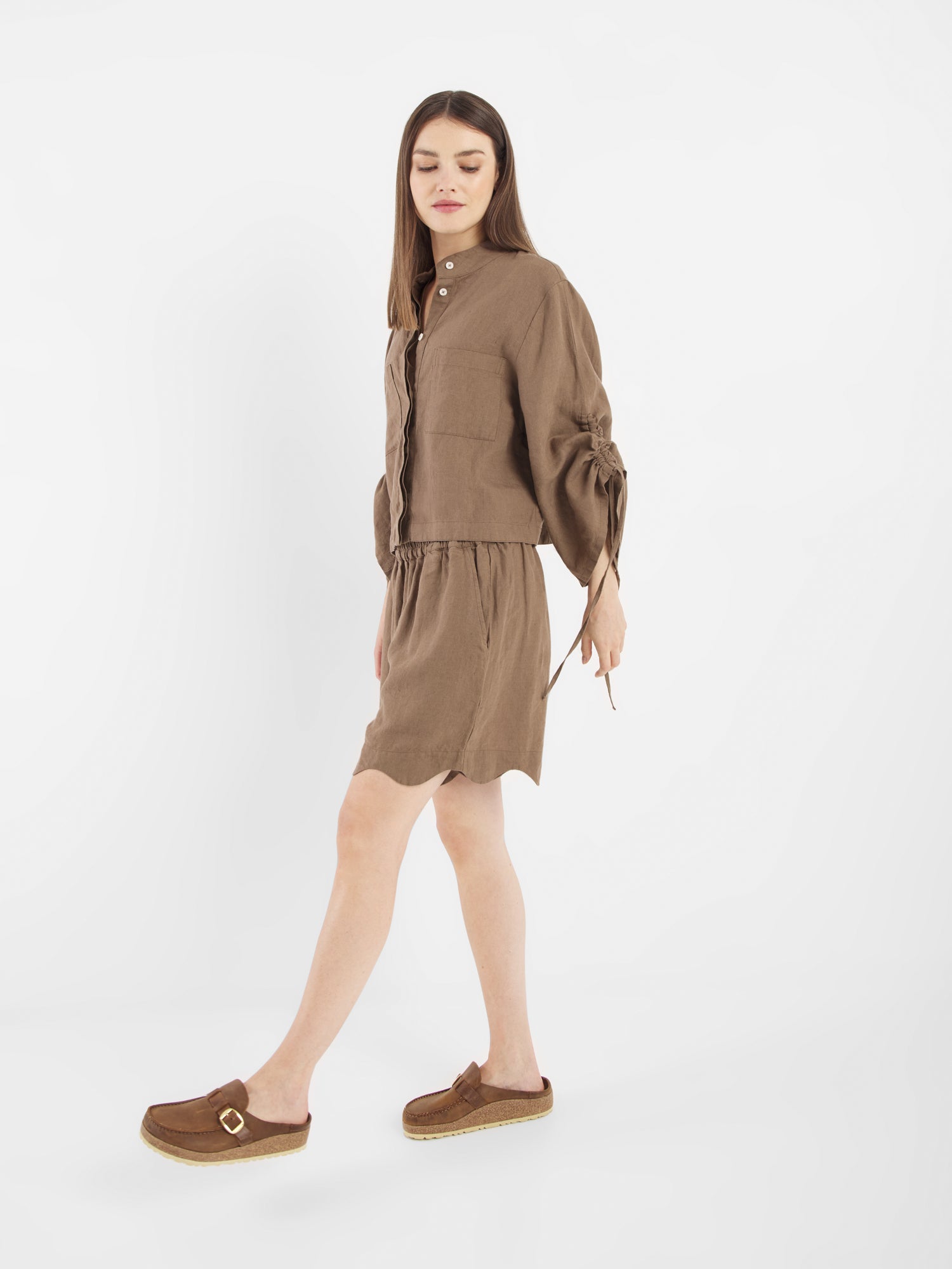 Eine Frau mit langen braunen Haaren trägt ein braunes, geknöpftes Kleid mit langen Ärmeln und Bindedetails über der Bluse Mary Linen Soft Brown, gepaart mit braunen Slippern, und läuft vor einem schlichten weißen Hintergrund.