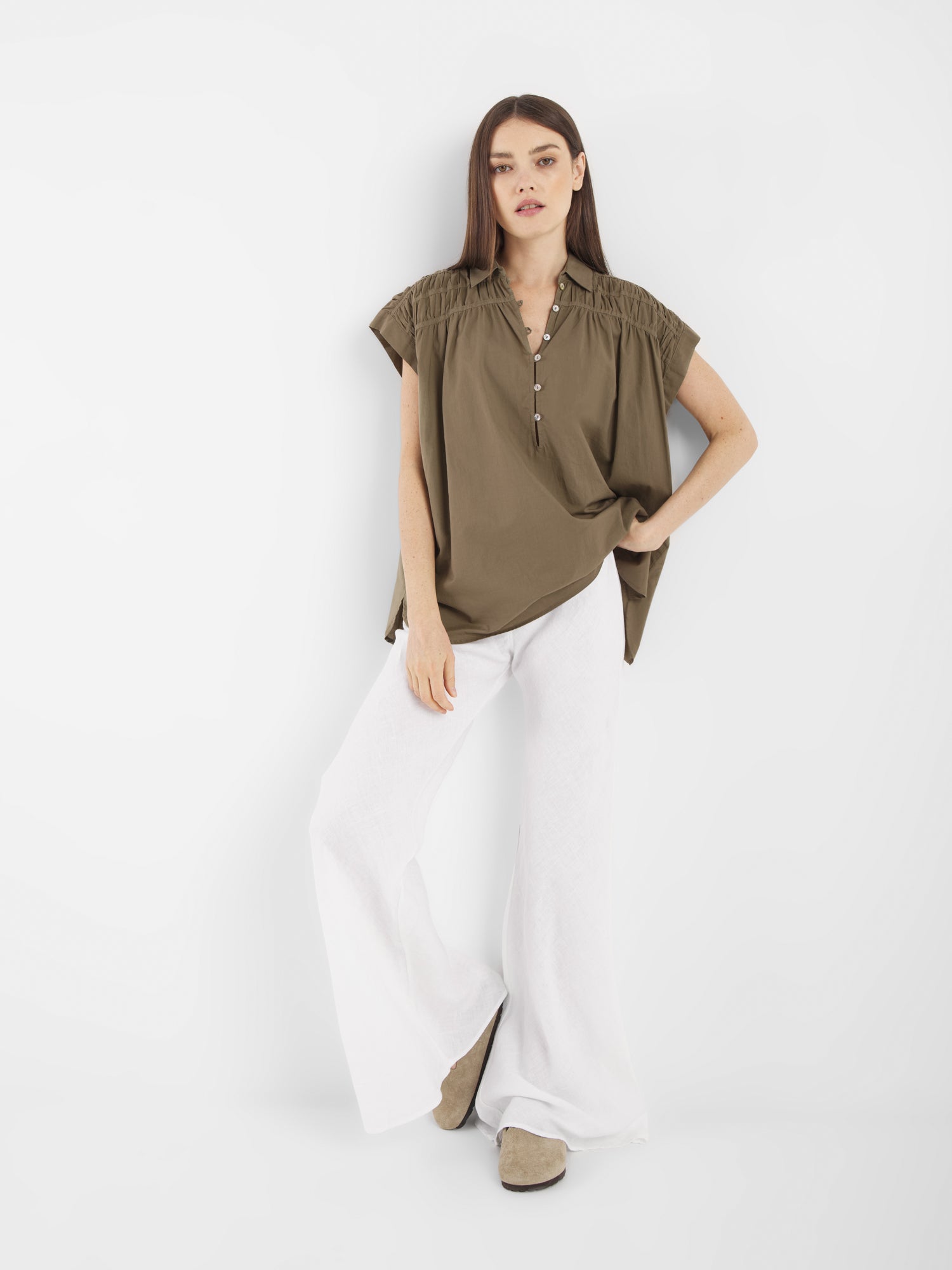 Eine Frau mit langem glattem Haar und neutralem Gesichtsausdruck posiert vor einem weißen Hintergrund und trägt die Bluse Maui Olive mit kurzen Ärmeln, gepaart mit einer weißen Hose mit weitem Bein und beigen Schuhen.