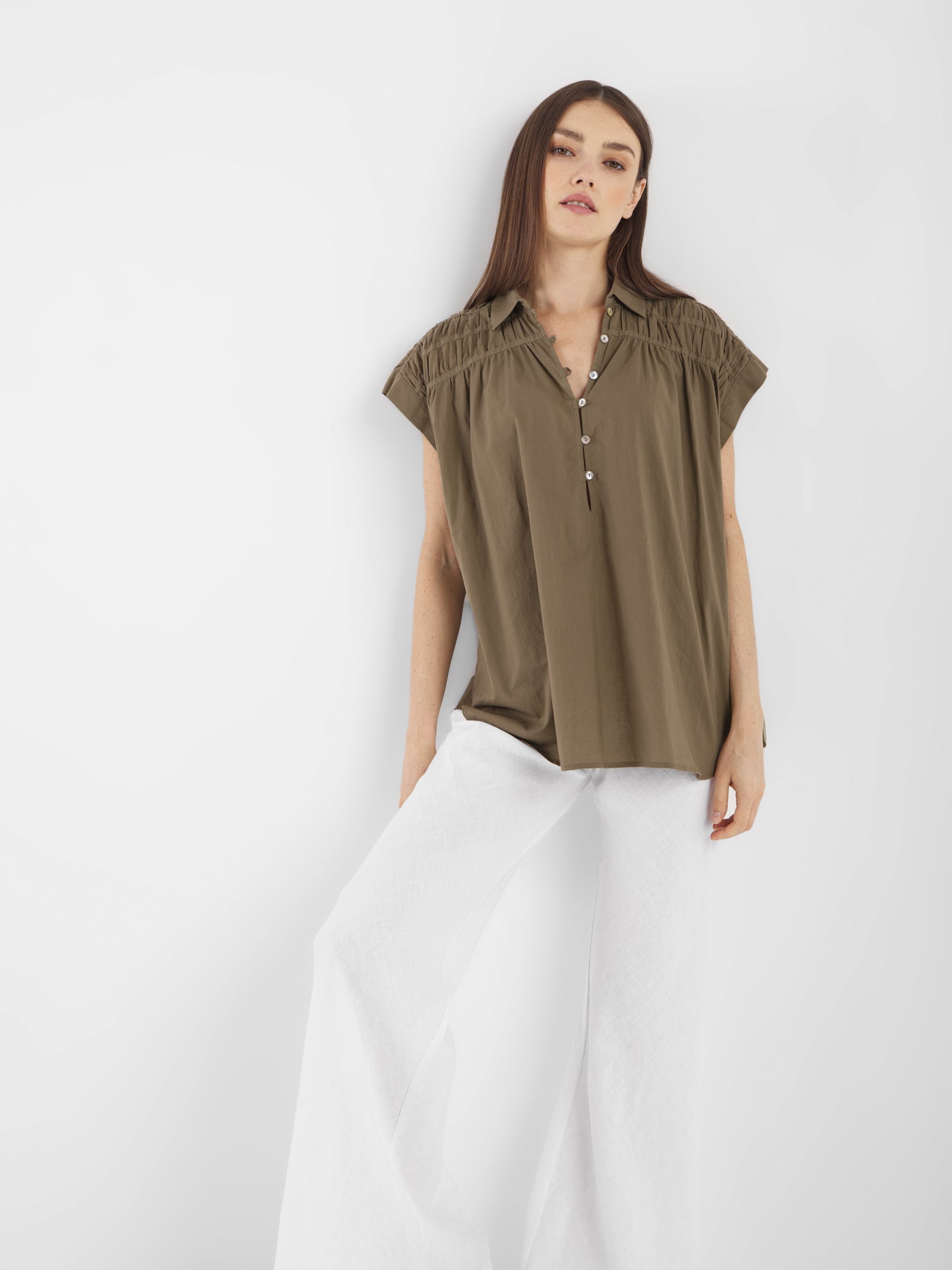 Eine Frau steht vor einem weißen Hintergrund und trägt die Bluse Maui Olive mit Knopfleiste und eine weite weiße Hose. Sie hat langes, glattes Haar und einen neutralen Ausdruck - die perfekte Sommerbluse für Frauen.