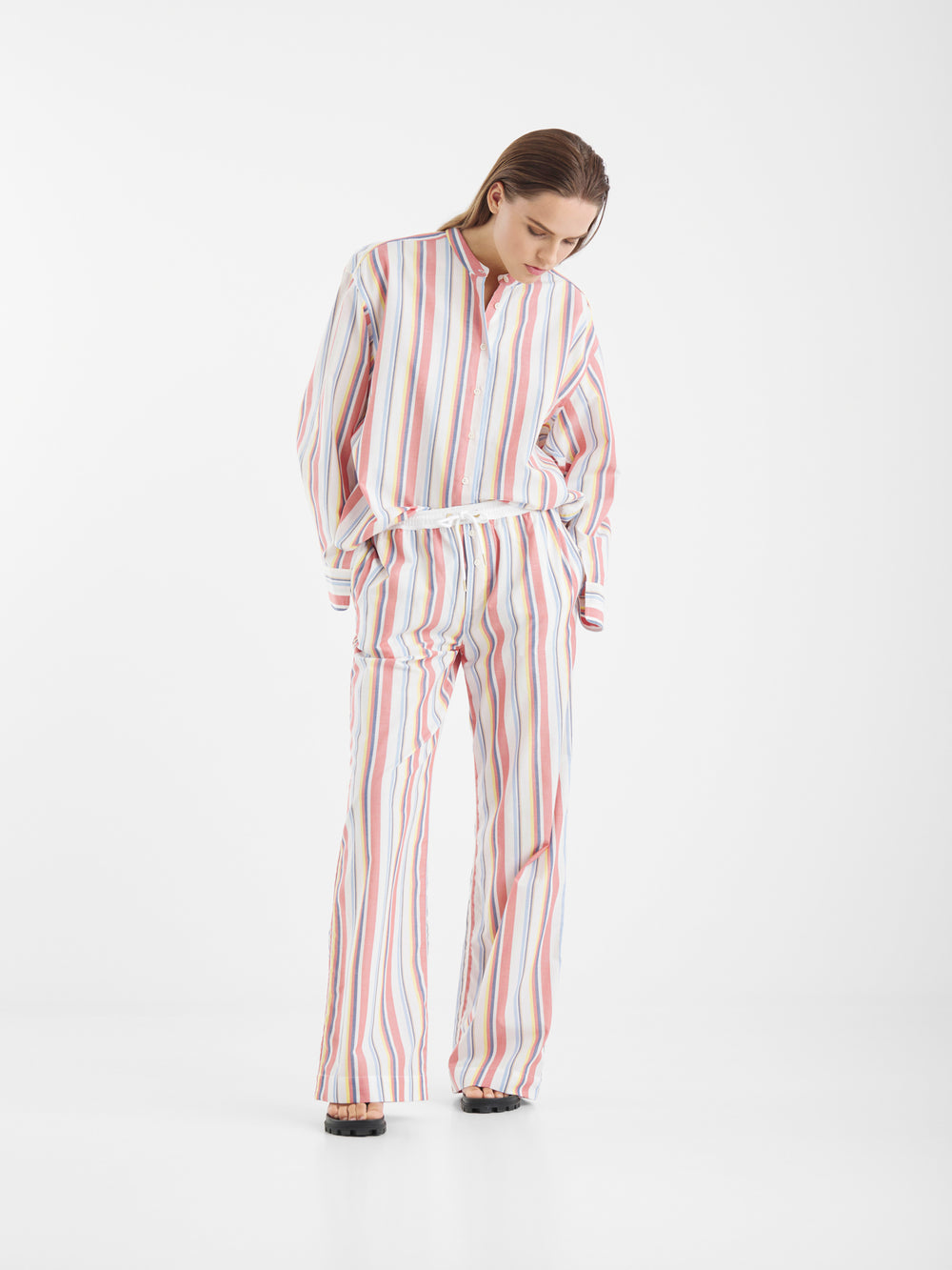Eine Person, die das Color Stripe Therapie Set trägt, posiert vor einem weißen Hintergrund in einem lockeren, langärmeligen Oberteil und einer Hose mit weiten Beinen, die mit auffälligen, farbigen vertikalen Streifen versehen sind, und schaut mit den Händen in den Taschen nach unten.