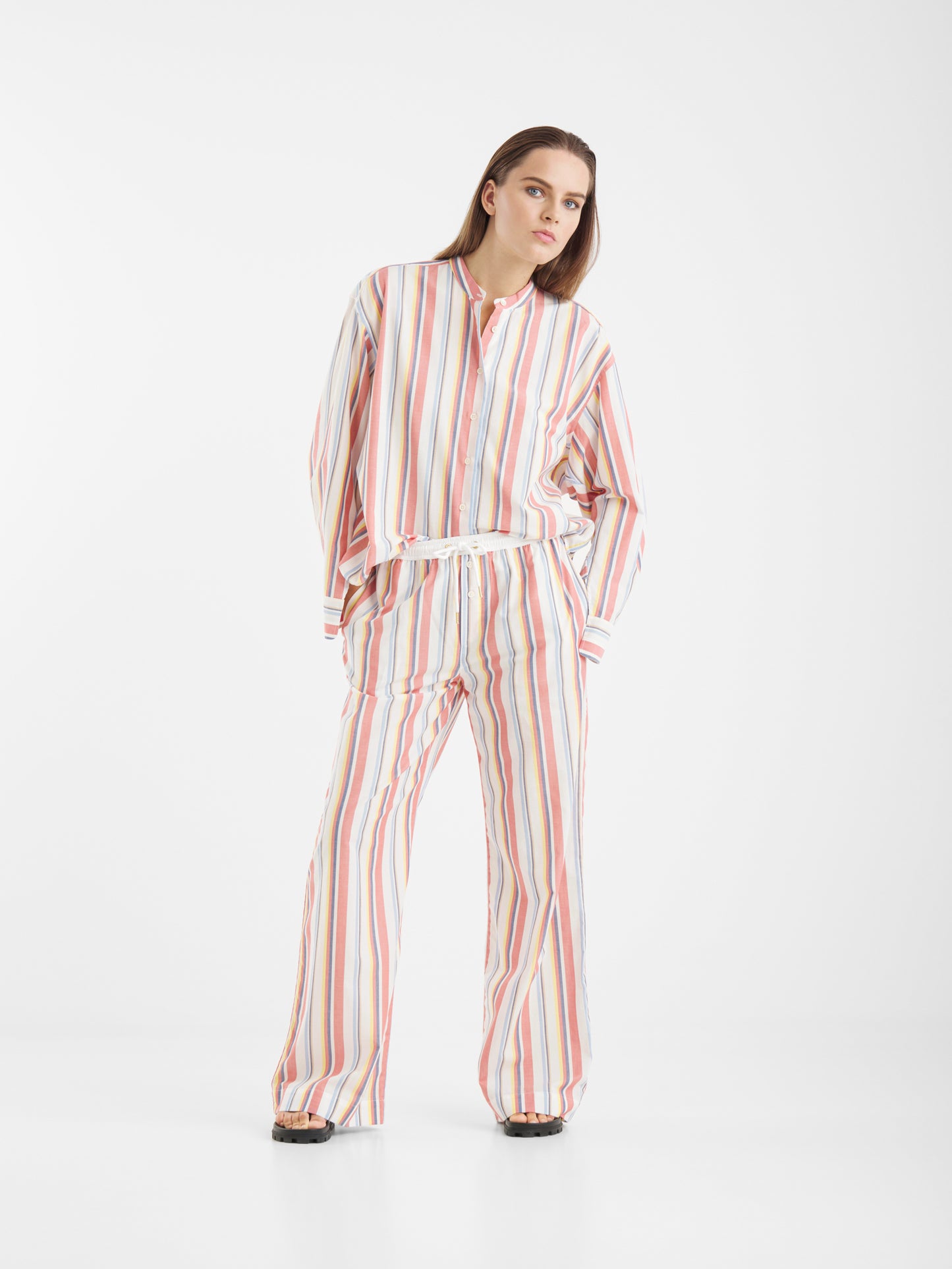Eine Frau steht vor einem weißen Hintergrund und trägt die Blouse New York Stripes, ein passendes Set mit einem locker sitzenden, langärmeligen Hemd und einer Hose mit weitem Bein, die vertikale Streifen in Pastellrosa, Orange, Blau und Weiß aufweist.