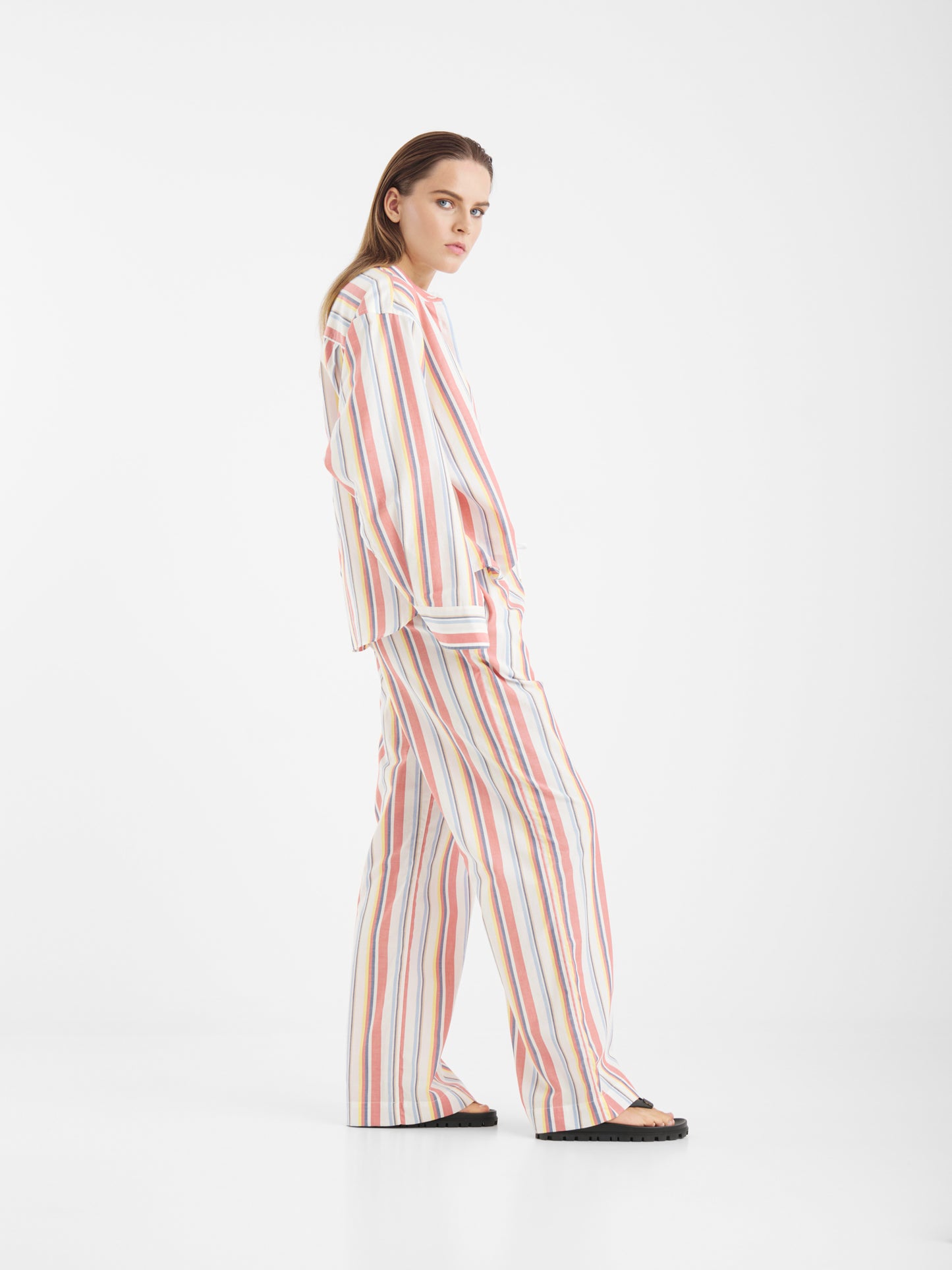 Eine Frau steht seitlich und schaut in die Kamera. Sie trägt die Bluse New York Stripes mit vertikalen pastellfarbenen Streifen und schwarzen Sandalen vor einem schlichten weißen Hintergrund.