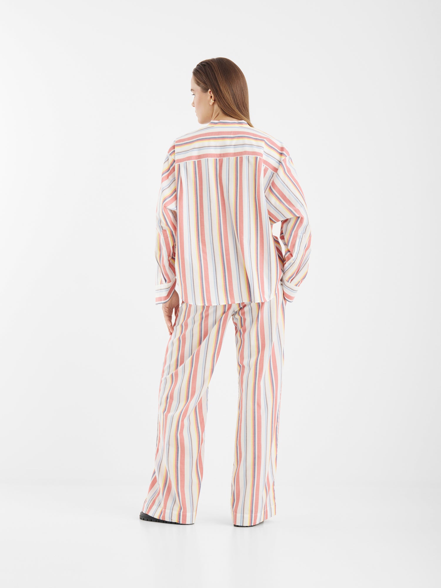 Eine Frau steht mit dem Rücken zur Kamera in einem schlichten weißen Studio und trägt die Blouse New York Stripes - ein lockeres, langärmeliges Oberteil mit mehrfarbigen vertikalen Streifen auf weißem Grund, gepaart mit einer passenden Hose mit weitem Bein.