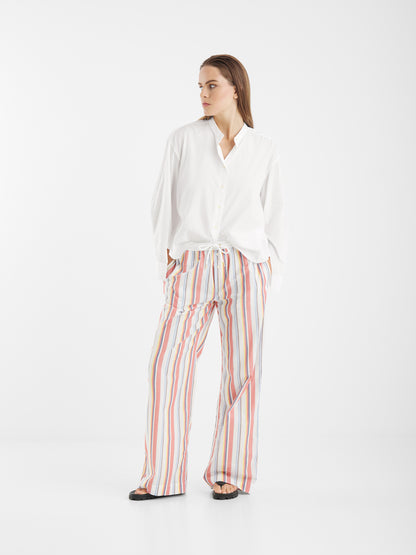 Eine Person steht vor einem schlichten weißen Hintergrund und trägt die Bluse New York White mit Statement-Ärmeln und eine bunte, vertikal gestreifte Hose mit Sandalen. Sie hat glattes braunes Haar und schaut mit den Händen in den Taschen zur Seite.