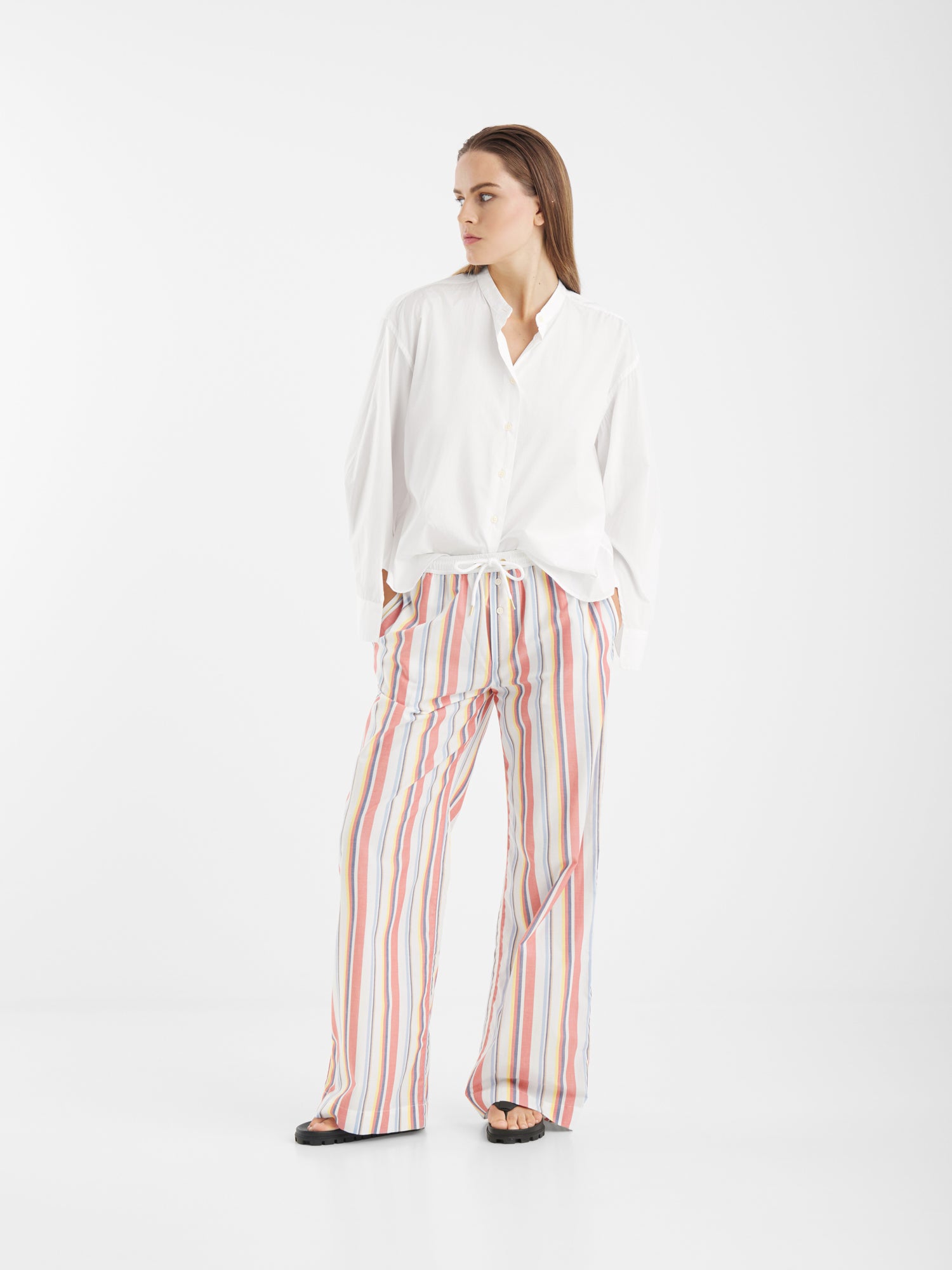 Eine Person steht vor einem einfarbig weißen Hintergrund, trägt die Bluse New York Stripes und eine lockere, mehrfarbige, vertikal gestreifte Hose mit schwarzen Sandalen und schaut mit den Händen in den Taschen zur Seite.