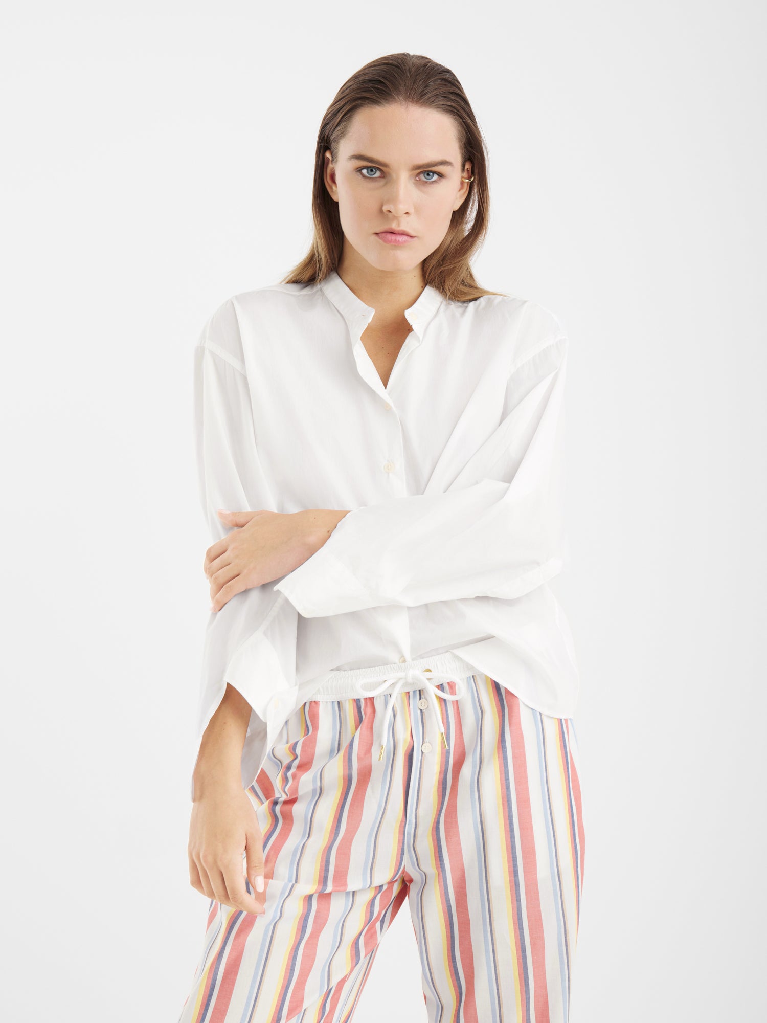 Eine Frau mit glattem braunem Haar trägt die Bluse New York White mit Statement-Ärmeln und eine bunt gestreifte Hose. Sie steht vor einem schlichten weißen Hintergrund, hat einen Arm gekreuzt und einen neutralen Ausdruck.