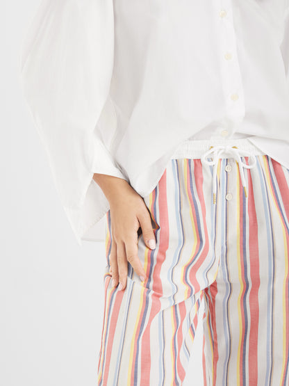 Eine Person trägt ein lockeres weißes Button-up-Hemd und eine bunt gestreifte Hose der Marke Pants New York mit Tunnelzug in der Taille, wobei sie eine Hand in die Hüfte stemmt.