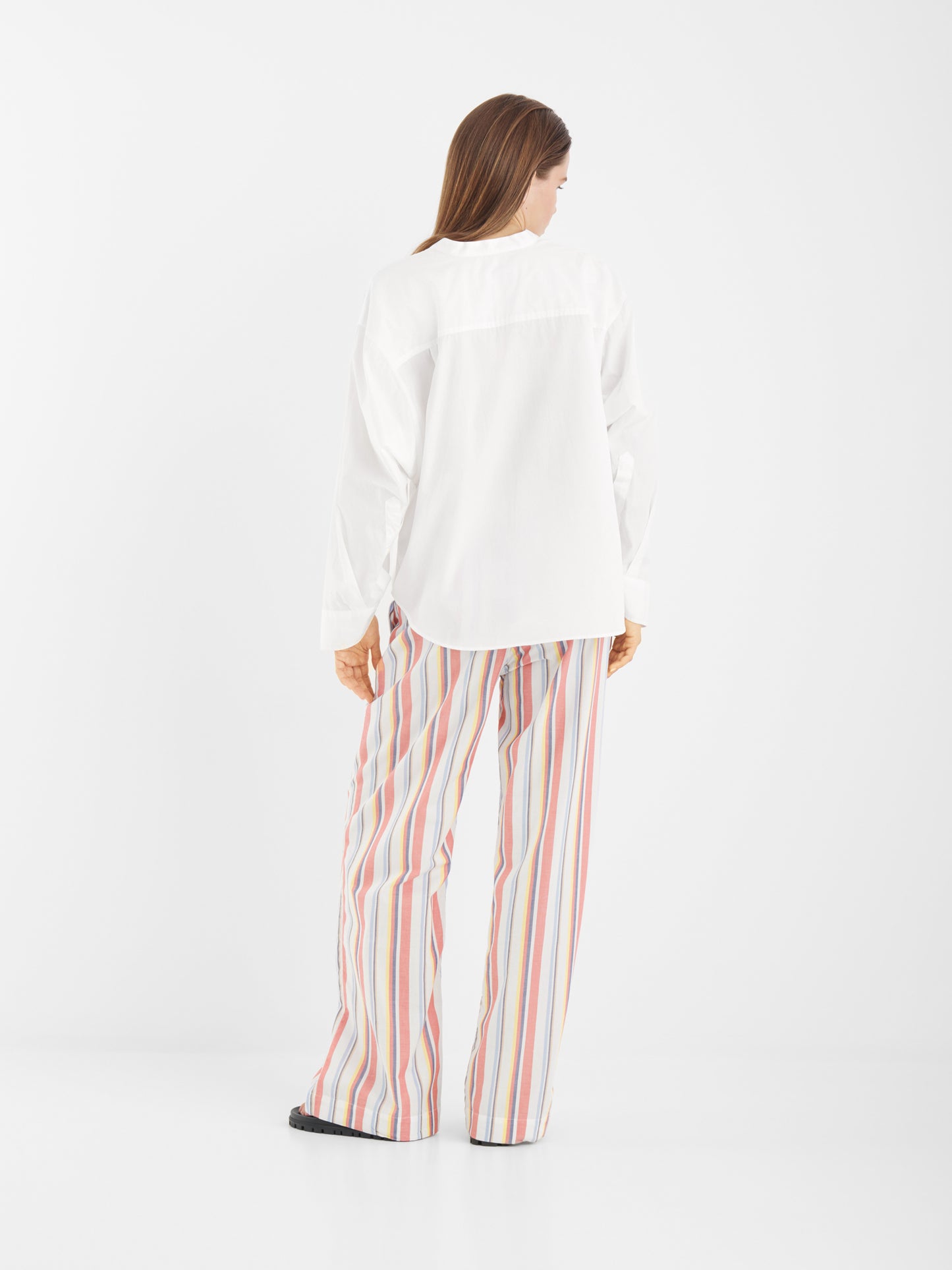 Eine Person steht mit dem Gesicht zur Wand und trägt die Bluse New York White mit Statement-Ärmeln und eine lockere, mehrfarbig vertikal gestreifte Hose vor einem schlichten weißen Hintergrund.