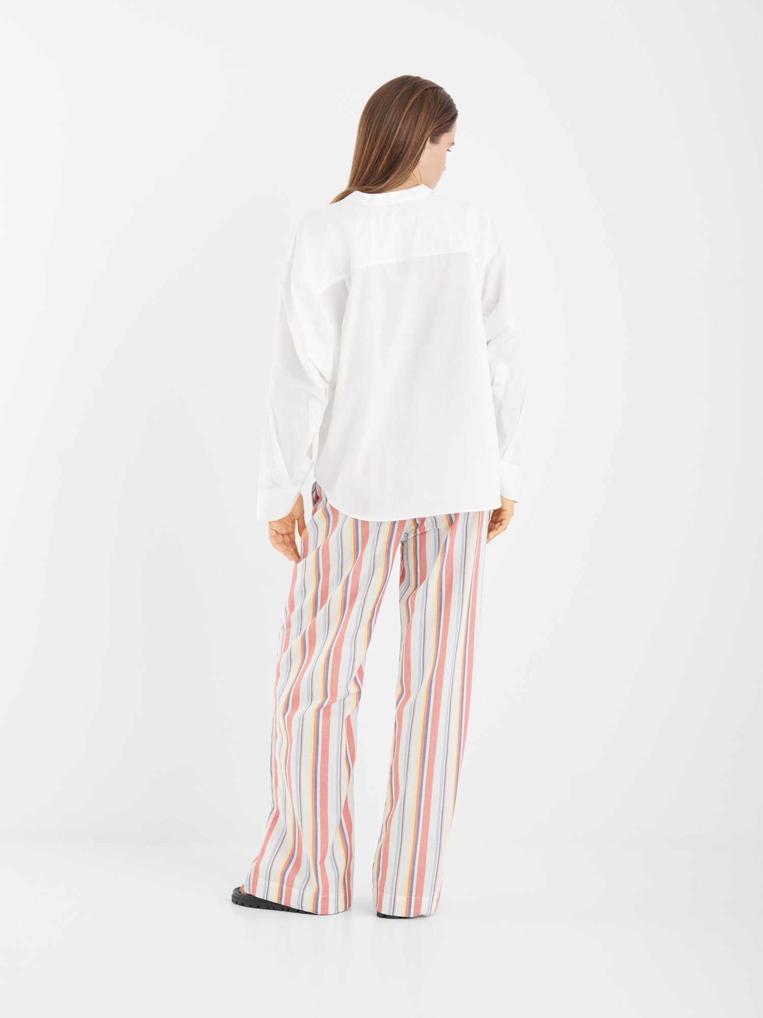 Eine Person steht mit dem Gesicht zur Wand und trägt die Bluse New York White mit Statement-Ärmeln und eine lockere, mehrfarbig vertikal gestreifte Hose vor einem schlichten weißen Hintergrund.