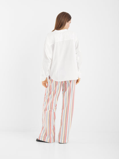 Eine Person steht mit dem Gesicht zur Wand und trägt die Bluse New York White mit Statement-Ärmeln und eine lockere, mehrfarbig vertikal gestreifte Hose vor einem schlichten weißen Hintergrund.