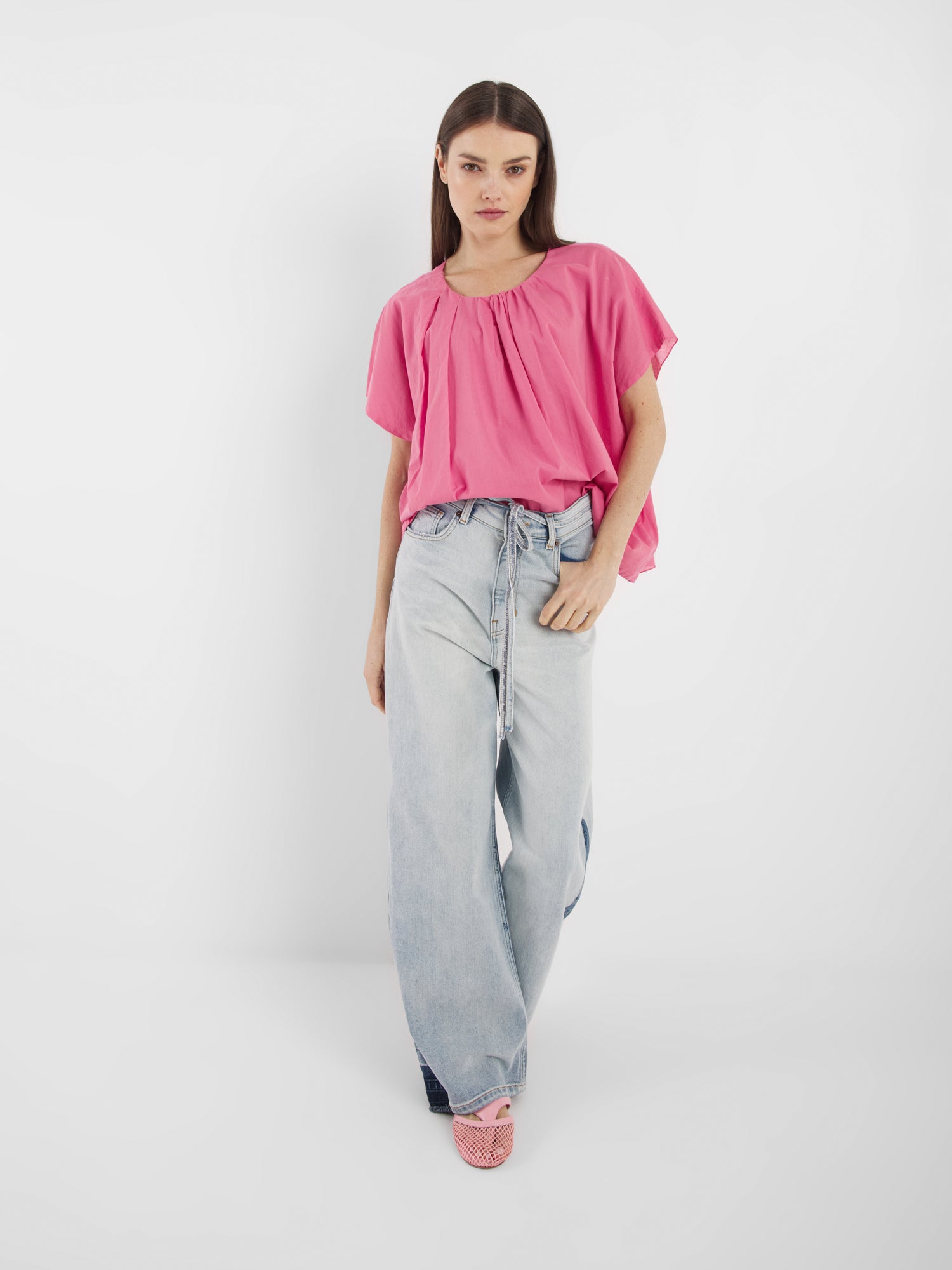 Eine Frau mit glattem braunem Haar und neutralem Gesichtsausdruck steht vor einem weißen Hintergrund und trägt die Bluse Tulip Pink, hellblaue Jeans mit weitem Bein und Schnürung sowie rosa Webschuhe.
