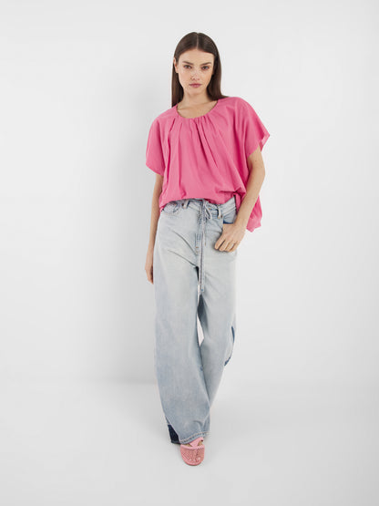 Eine Frau mit glattem braunem Haar und neutralem Gesichtsausdruck steht vor einem weißen Hintergrund und trägt die Bluse Tulip Pink, hellblaue Jeans mit weitem Bein und Schnürung sowie rosa Webschuhe.