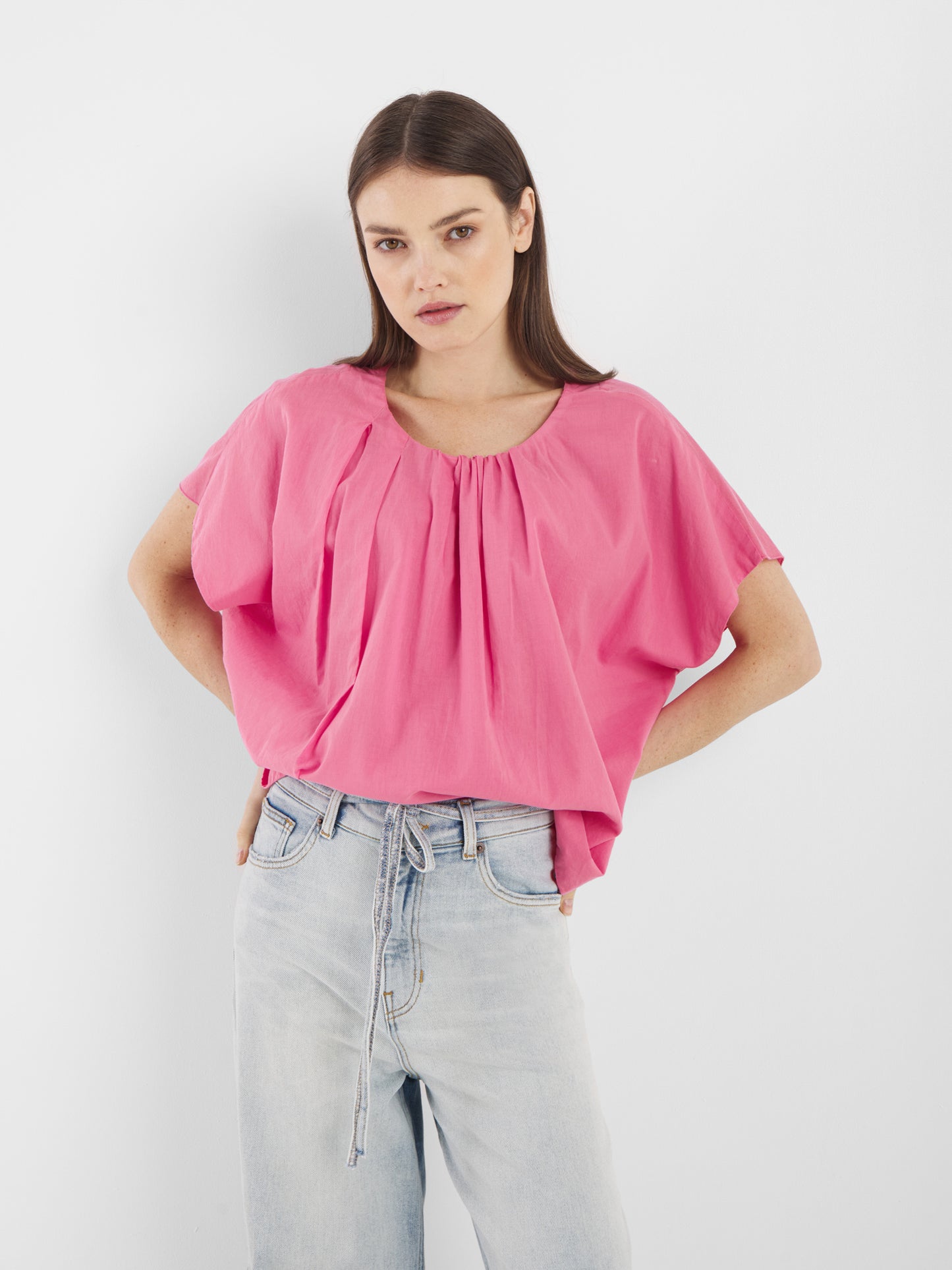 Eine Frau mit glattem, dunkelbraunem Haar und neutralem Gesichtsausdruck steht vor einem weißen Hintergrund. Sie trägt die Bluse Tulip Pink mit kurzen Ärmeln und lockerer Passform, gepaart mit einer hellen, hoch taillierten Jeans mit Bindegürtel.
