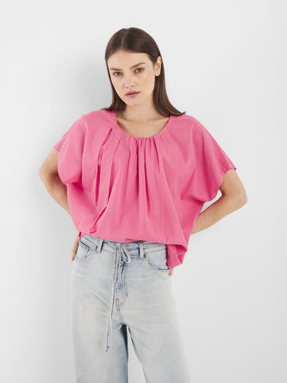 Eine Frau mit glattem, dunkelbraunem Haar und neutralem Gesichtsausdruck steht vor einem weißen Hintergrund. Sie trägt die Bluse Tulip Pink mit kurzen Ärmeln und lockerer Passform, gepaart mit einer hellen, hoch taillierten Jeans mit Bindegürtel.