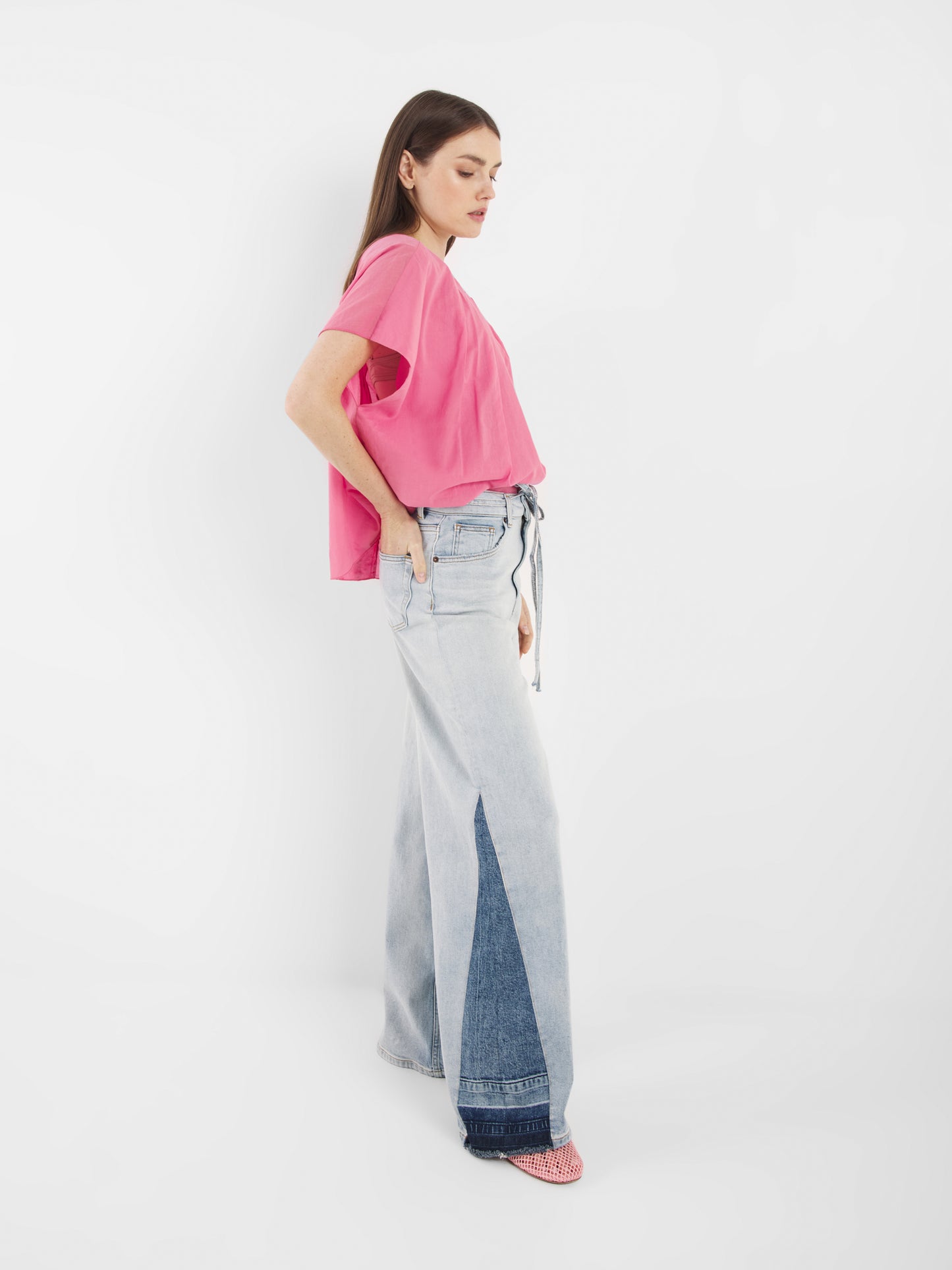 Eine Frau im Profil vor weißem Hintergrund trägt die Bluse Tulip Pink, ein hellrosa, kurzärmeliges Oberteil, gepaart mit einer hellblauen Jeans mit weitem Bein und einem dunkelblauen, dreieckigen Denim-Einsatz. Sie hat langes braunes Haar und rosa Schuhe.