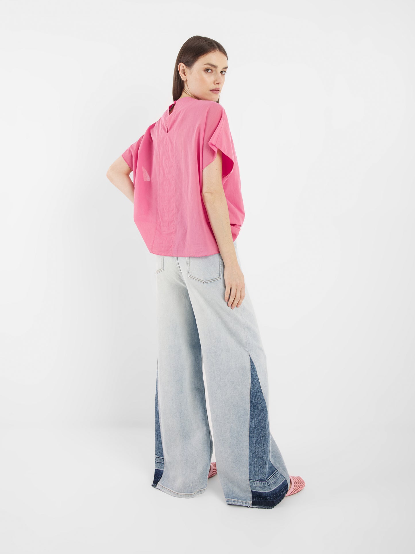 Eine Frau steht in einem Atelier und trägt die Bluse Tulip Pink mit hellen Jeans mit weitem Bein und dunklen Jeanseinsätzen. Sie blickt über ihre Schulter, hat glattes braunes Haar und rosa Schuhe.