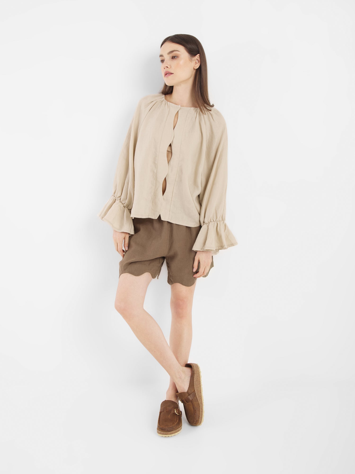 Eine Frau steht vor einem weißen Hintergrund und trägt die Bluse Wave Linen Beige mit gerüschten Manschetten, gepaart mit braunen Shorts und Slippern. Sie blickt leicht zur Seite und hat ein Bein über das andere geschlagen.
