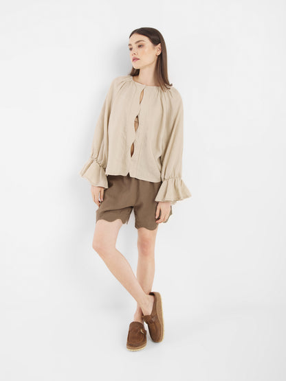 Eine Frau steht vor einem weißen Hintergrund und trägt die Bluse Wave Linen Beige mit gerüschten Manschetten, gepaart mit braunen Shorts und Slippern. Sie blickt leicht zur Seite und hat ein Bein über das andere geschlagen.