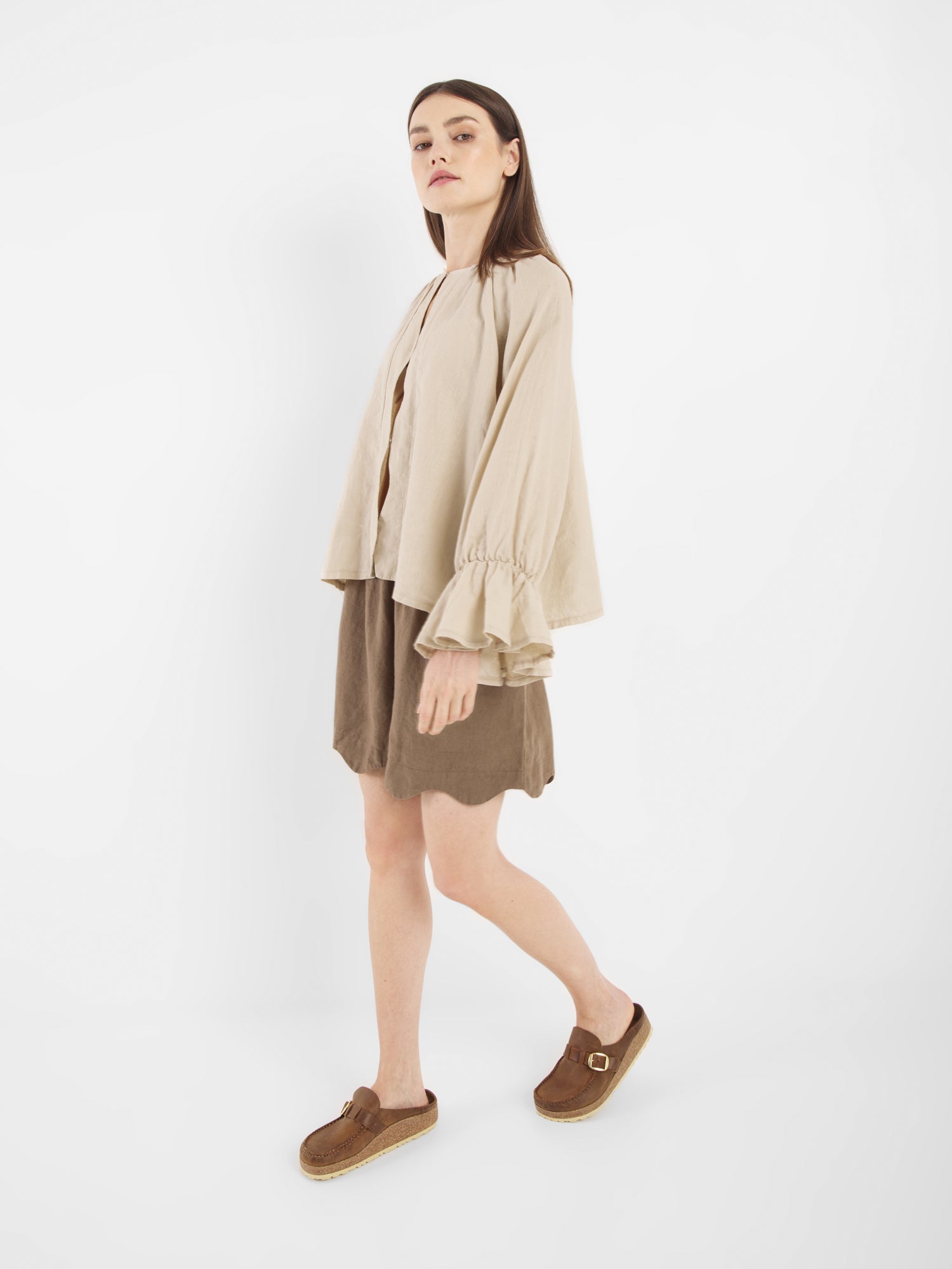 Eine Frau steht vor einem weißen Hintergrund und trägt die Bluse Wave Linen Beige. Sie blickt mit einem leicht angewinkelten Bein und neutraler Miene nach vorne und kombiniert das fließende, langärmelige Oberteil mit einem braunen Kleid und braunen Slippern.