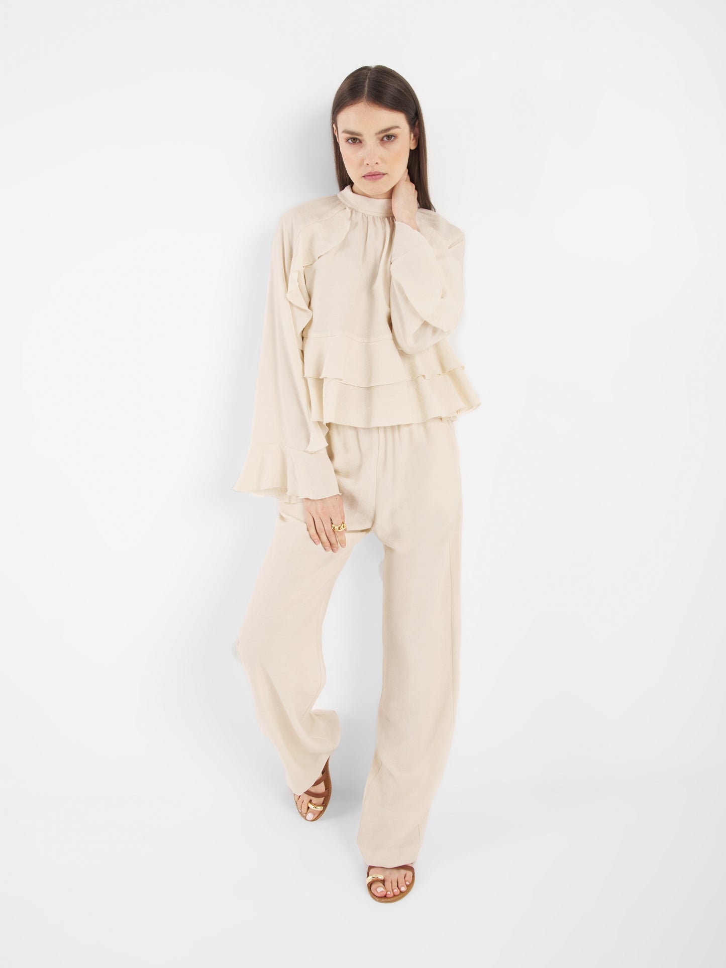 Eine Frau mit glattem braunem Haar und neutralem Gesichtsausdruck steht vor einem weißen Hintergrund. Sie trägt die Bluse Zoya Off white mit Rüschendetails und eine Hose mit weitem Bein, zu der sie braune Sandalen trägt.