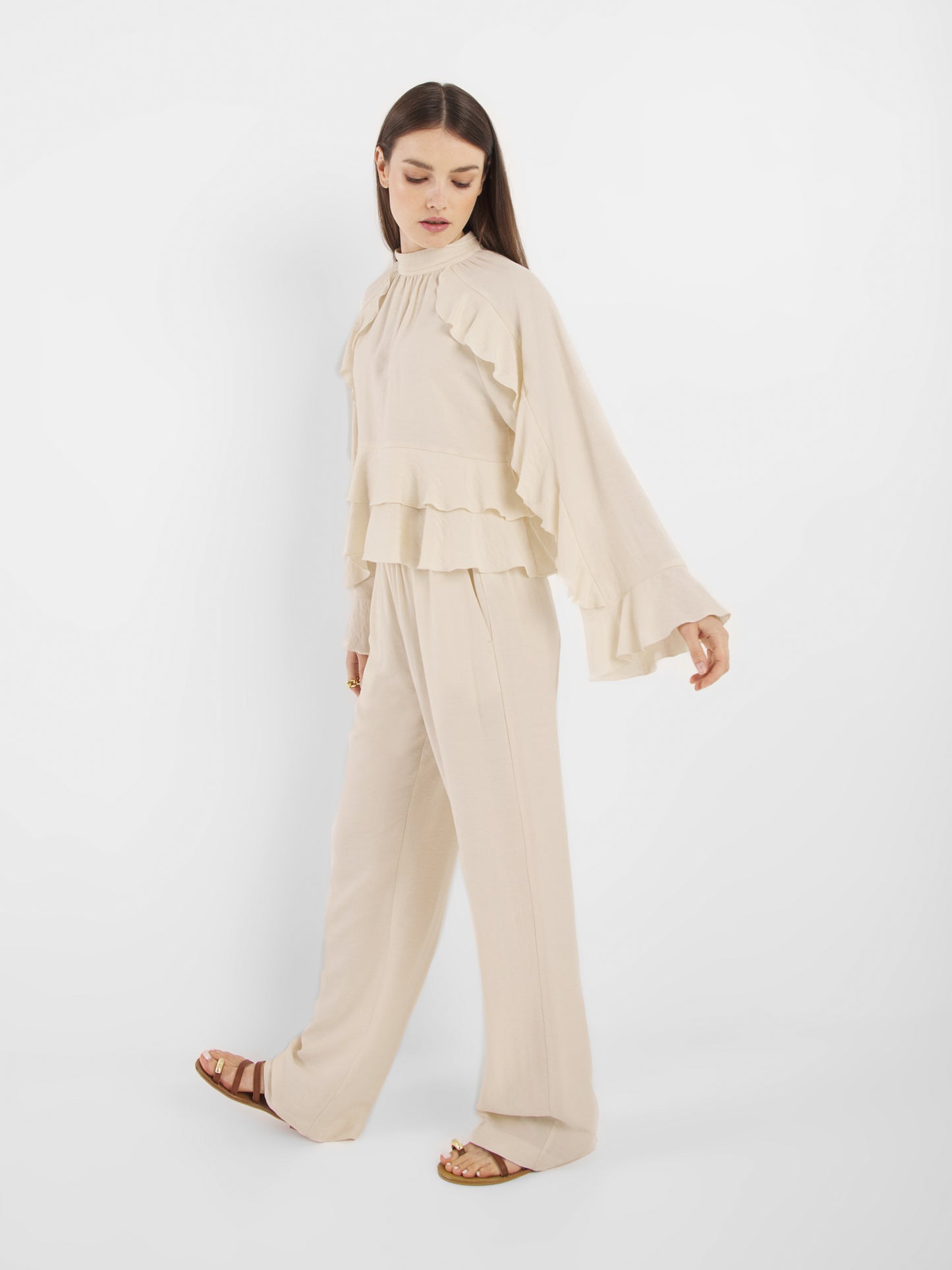 Eine Frau steht vor einem einfarbig weißen Hintergrund und trägt die Hose Draw Off White - eine locker sitzende, fließende Hose, die mit einem langärmeligen, gerüschten Oberteil kombiniert ist - und schaut nach unten.