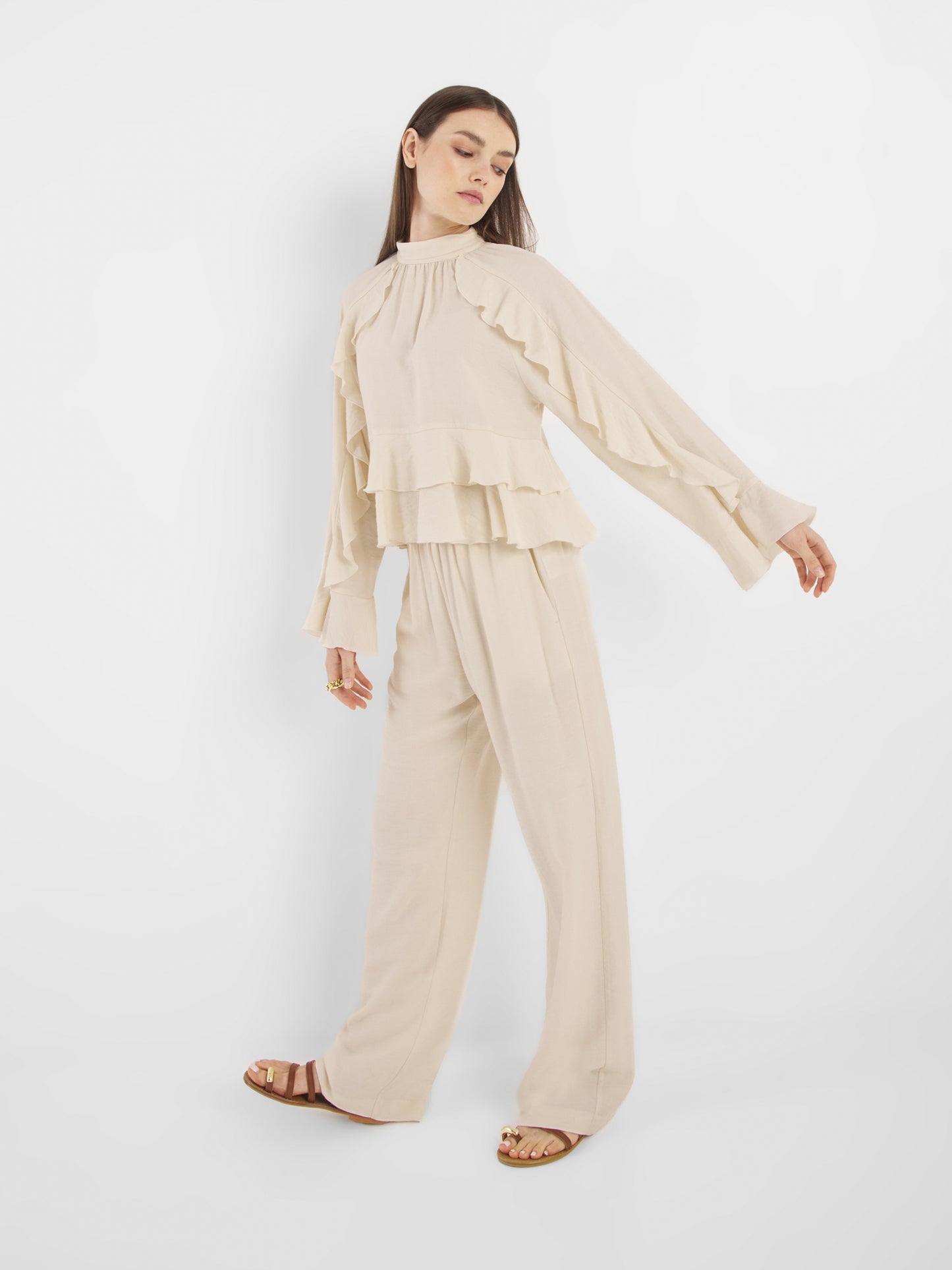 Eine Frau posiert vor einem weißen Hintergrund und trägt die Bluse Zoya Off White mit Rüschendetails, kombiniert mit einer Hose mit weitem Bein, braunen Sandalen und Goldringen.