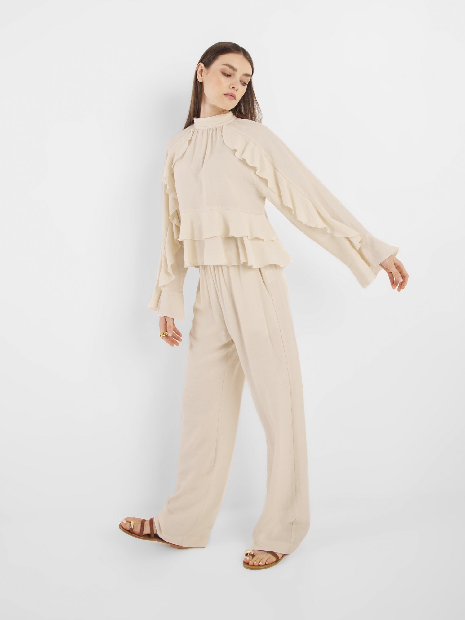Eine Frau posiert vor einem weißen Hintergrund und trägt die Bluse Zoya Off White mit Rüschendetails, kombiniert mit einer Hose mit weitem Bein, braunen Sandalen und Goldringen.