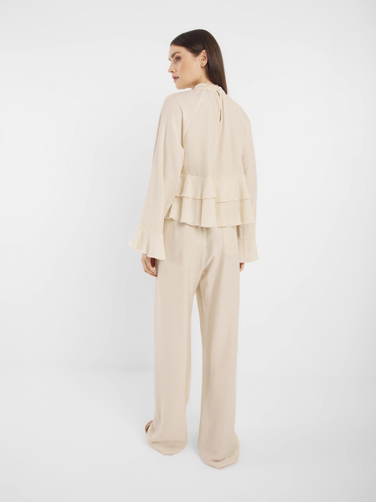 Die Frau steht mit dem Gesicht zur Wand und trägt ein hellbeiges, langärmeliges Oberteil mit mehrlagigen Rüschen am Rücken und eine Hose in Off-White, die sich vor einem schlichten weißen Hintergrund abzeichnet.