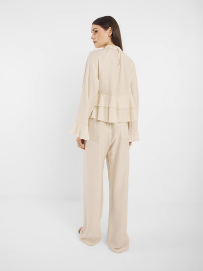 Die Frau steht mit dem Gesicht zur Wand und trägt ein hellbeiges, langärmeliges Oberteil mit mehrlagigen Rüschen am Rücken und eine Hose in Off-White, die sich vor einem schlichten weißen Hintergrund abzeichnet.