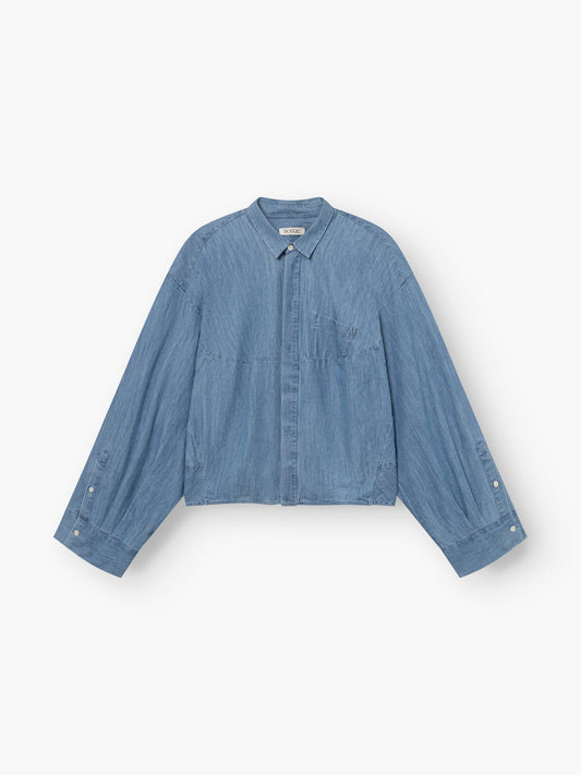 Das Blouse Bomber Denim ist ein hellblaues, langärmeliges Jeanshemd mit Kragen, geknöpfter Vorderseite, Brusttasche und geknöpften Manschetten, das flach auf weißem Hintergrund abgebildet ist.