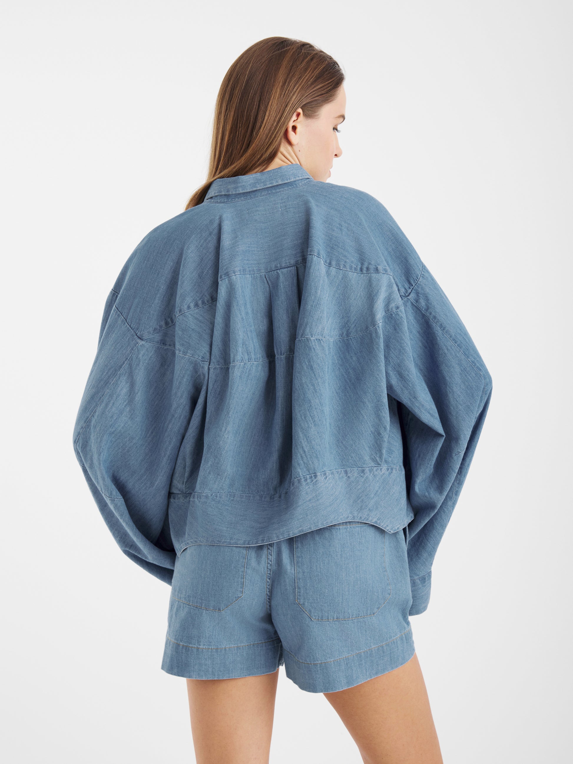 Eine Frau mit langen braunen Haaren, von hinten gesehen, trägt die Blouse Bomber Denim Jacke und dazu passende Jeansshorts vor einem schlichten weißen Hintergrund.