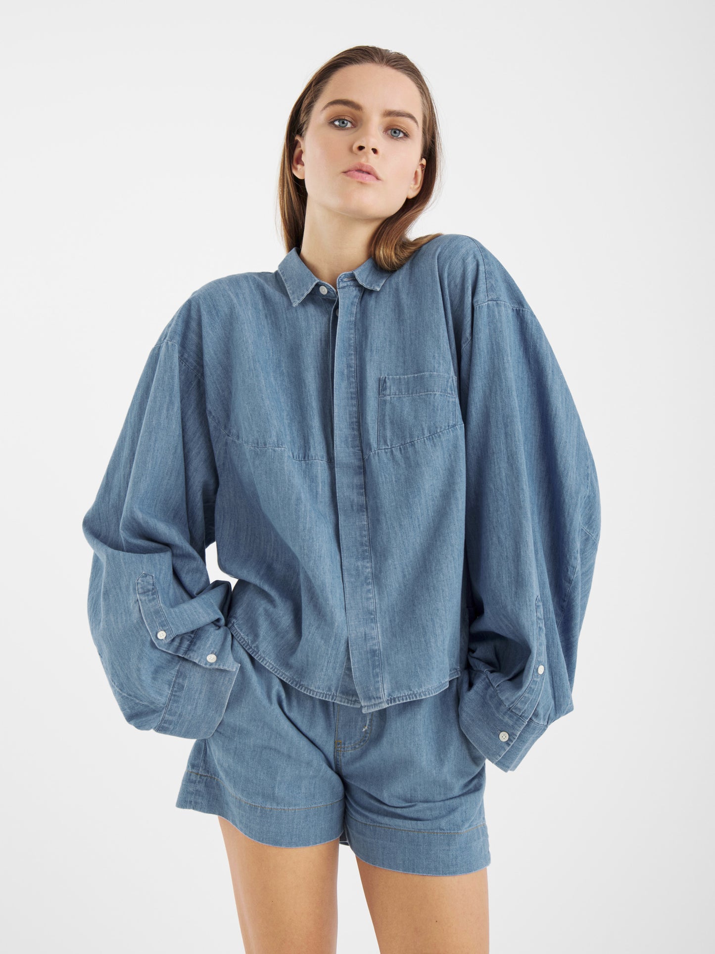 Eine Frau steht selbstbewusst vor einem weißen Hintergrund, die Hände in den Taschen, und trägt die Blouse Bomber Denim - ein übergroßes, langärmeliges Jeanshemd, das mit einer passenden kurzen Hose kombiniert wird.
