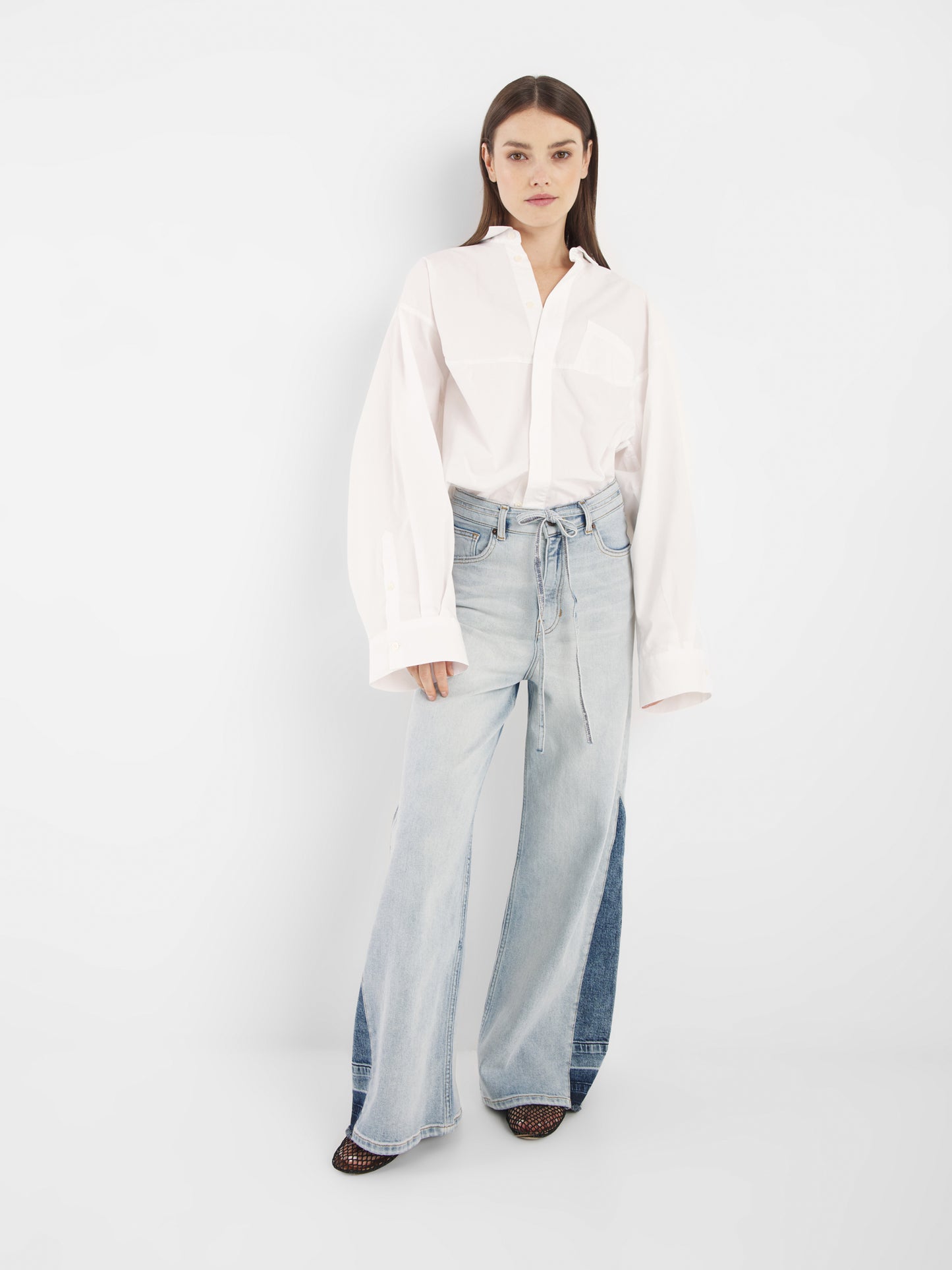 Eine Frau steht vor einem weißen Hintergrund und trägt den Blouse Bomber White, ein übergroßes, langärmeliges Button-up-Hemd, gepaart mit hellblauen, weit geschnittenen Jeans und spitzen Schuhen.