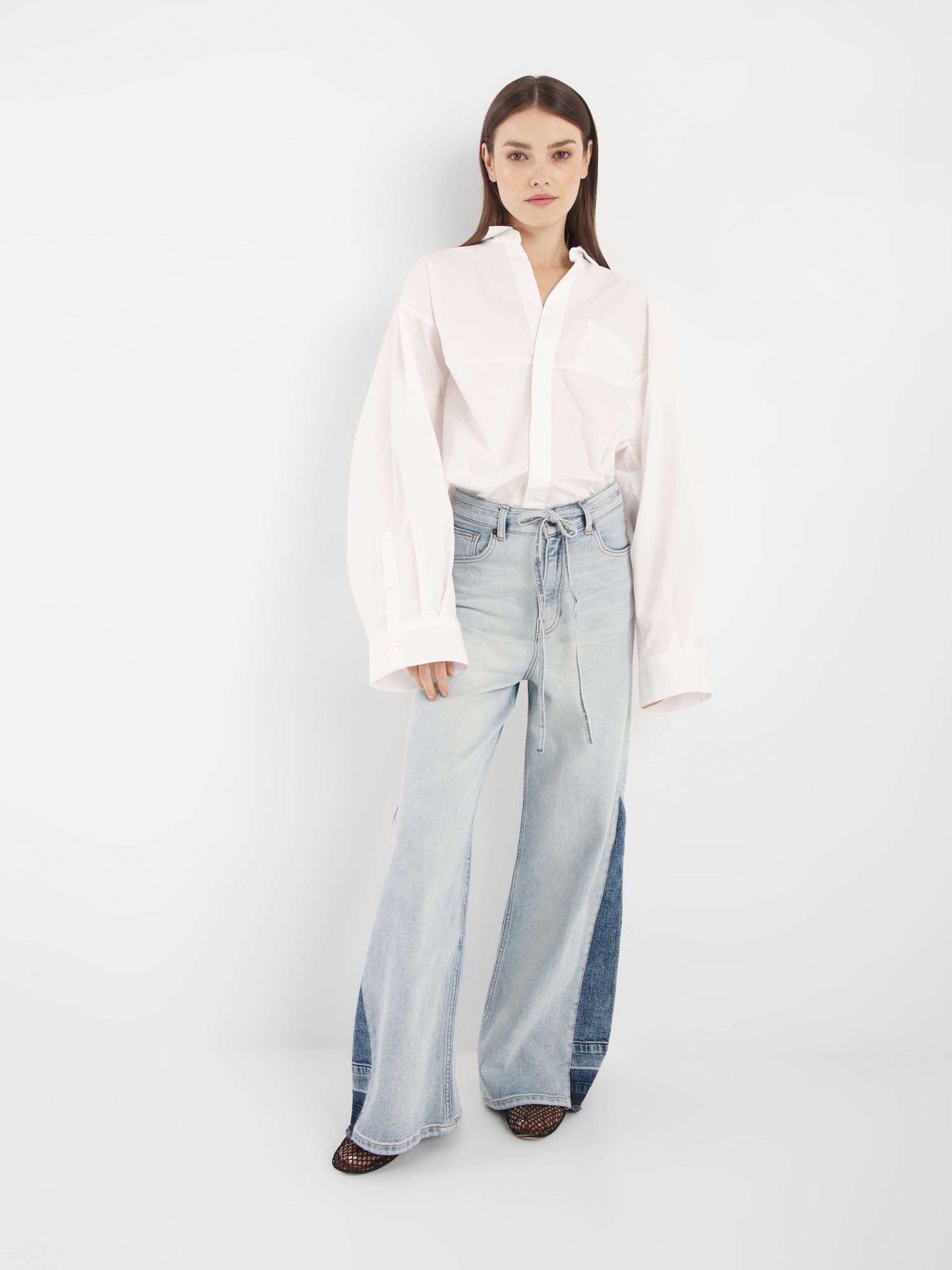 Eine Frau steht vor einem weißen Hintergrund und trägt den Blouse Bomber White, ein übergroßes, langärmeliges Button-up-Hemd, gepaart mit hellblauen, weit geschnittenen Jeans und spitzen Schuhen.