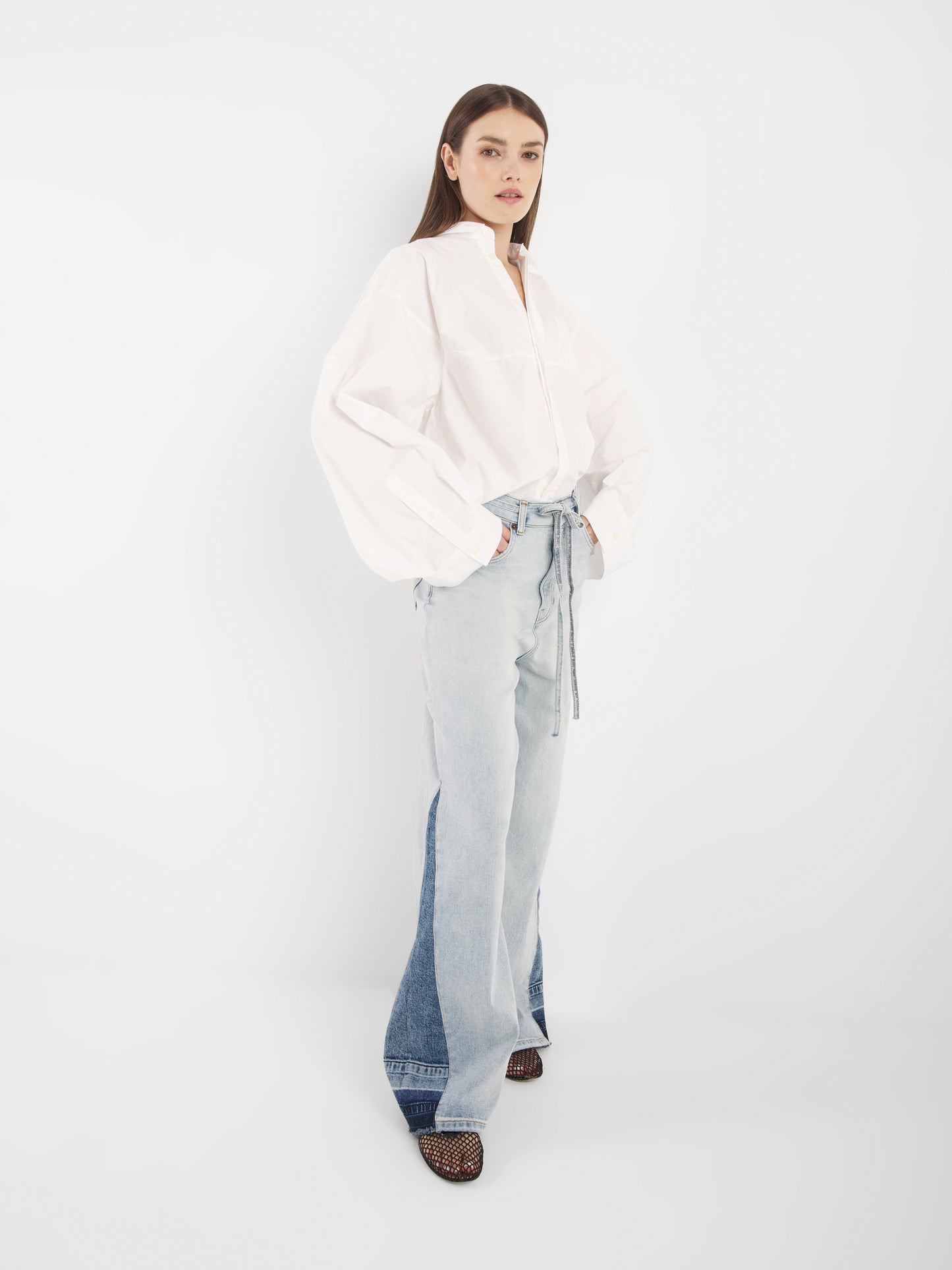 Eine Frau mit glattem braunem Haar und neutralem Gesichtsausdruck steht vor einem weißen Hintergrund und trägt den Blouse Bomber White, hellblaue Jeans mit hoher Taille und weitem Bein sowie schwarze offene Schuhe.