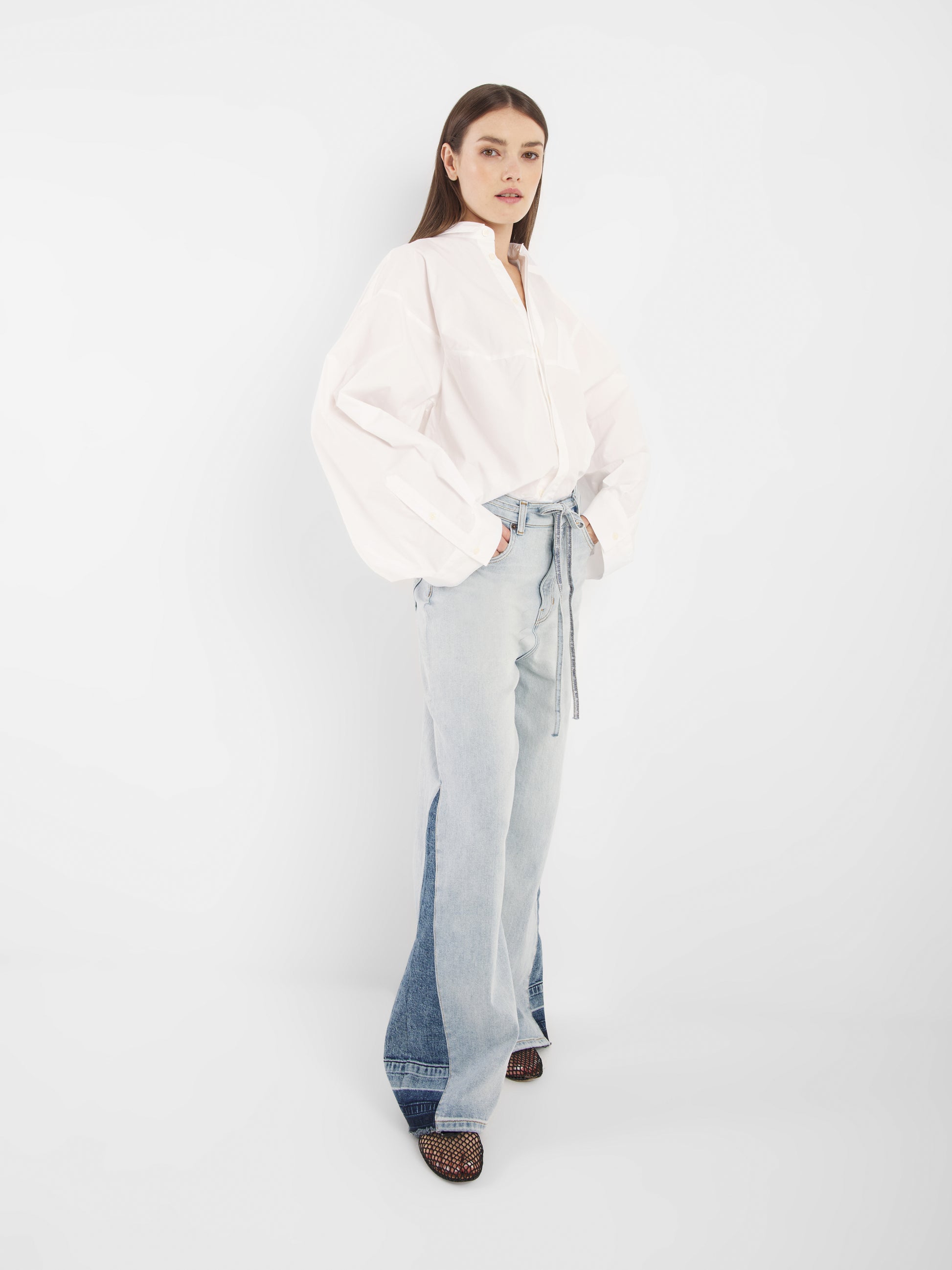 Eine Frau mit glattem braunem Haar und neutralem Gesichtsausdruck steht vor einem weißen Hintergrund und trägt den Blouse Bomber White, hellblaue Jeans mit hoher Taille und weitem Bein sowie schwarze offene Schuhe.