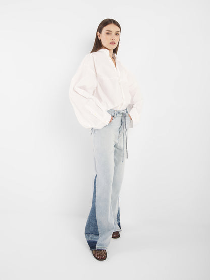 Eine Frau mit glattem braunem Haar und neutralem Gesichtsausdruck steht vor einem weißen Hintergrund und trägt den Blouse Bomber White, hellblaue Jeans mit hoher Taille und weitem Bein sowie schwarze offene Schuhe.