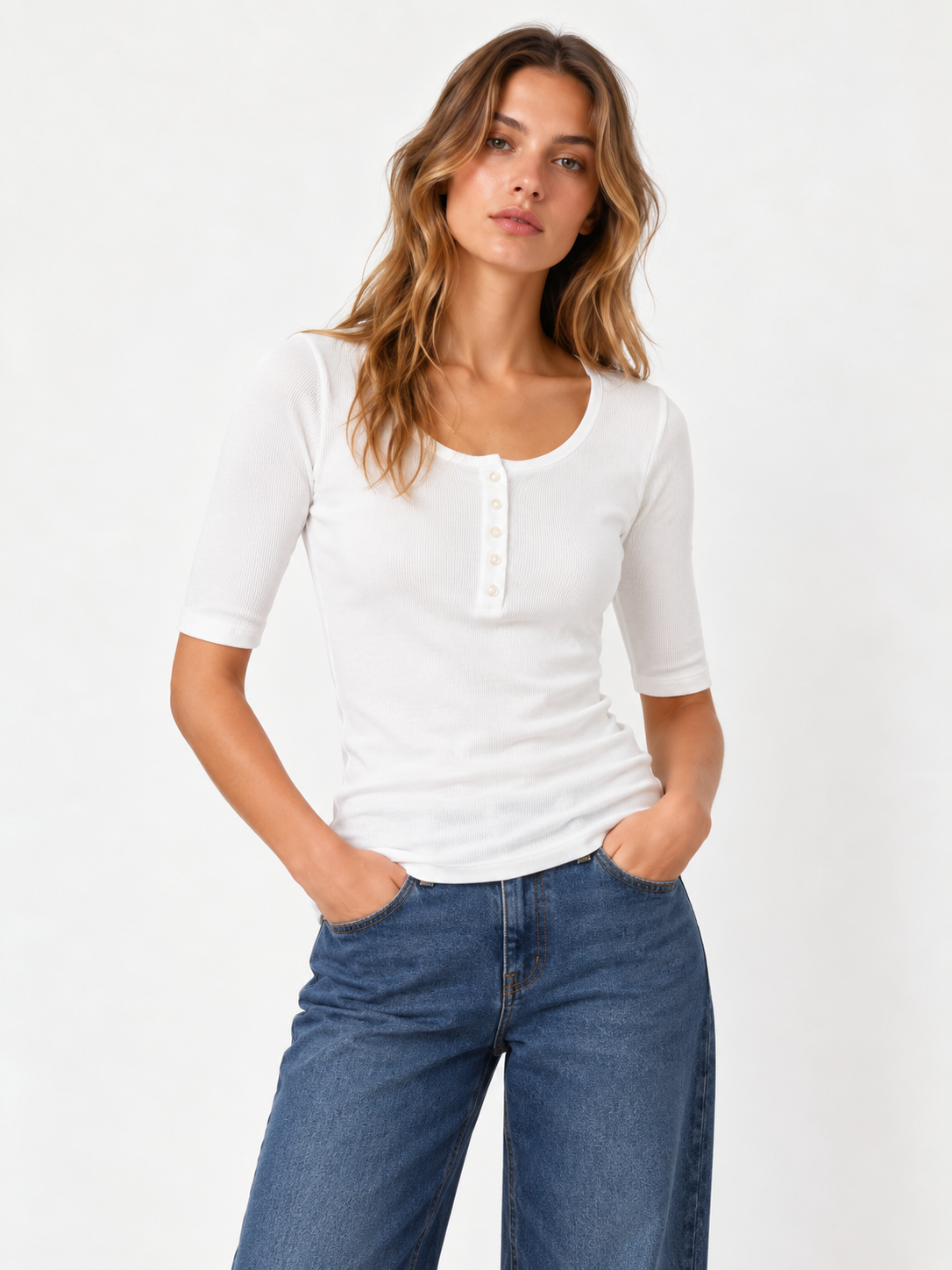 Eine Frau trägt das Modell Half Sleeve Button Rib White mit ellbogenlangen Ärmeln und eine blaue Jeans; sie steht mit den Händen in den Taschen vor einem einfarbigen Hintergrund.